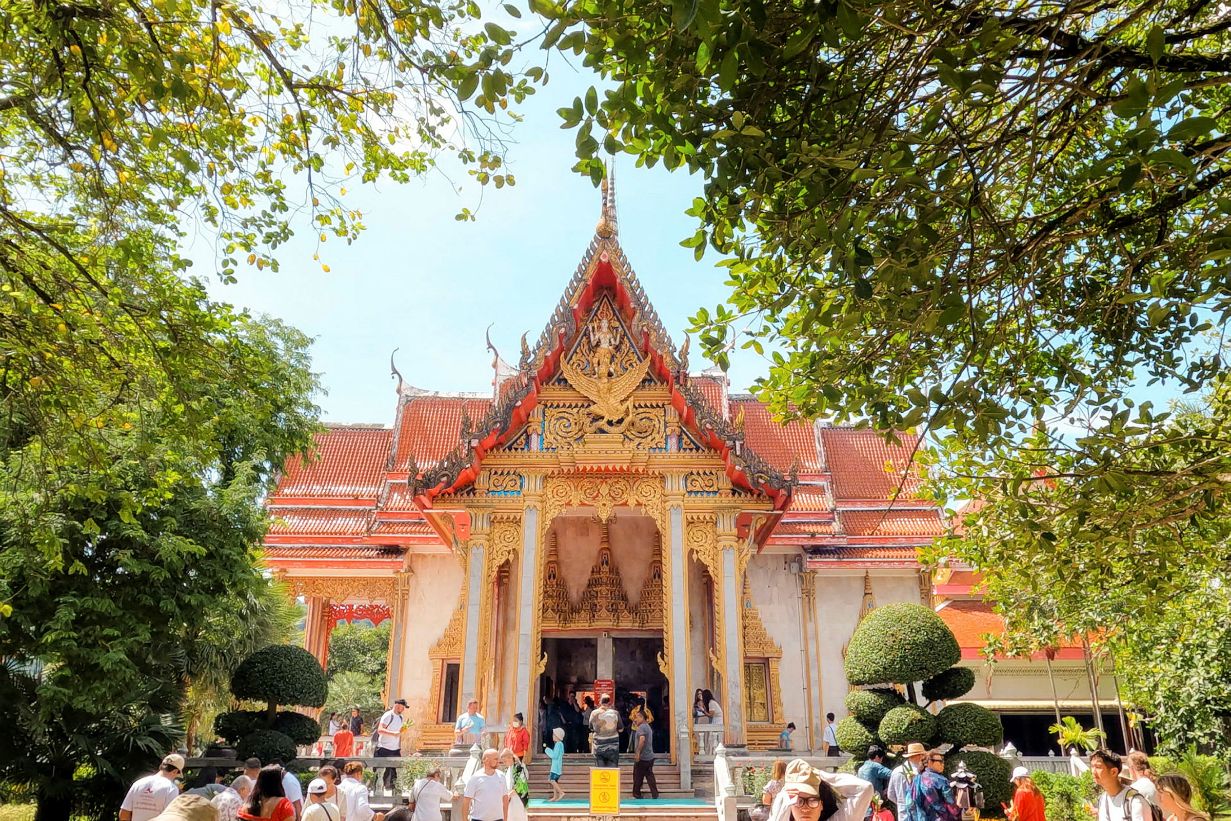 Billede af et detaljerigt thailandsk tempel omgivet af frodig vegetation, med besøgende foran bygningen. (AI-genereret tekst)