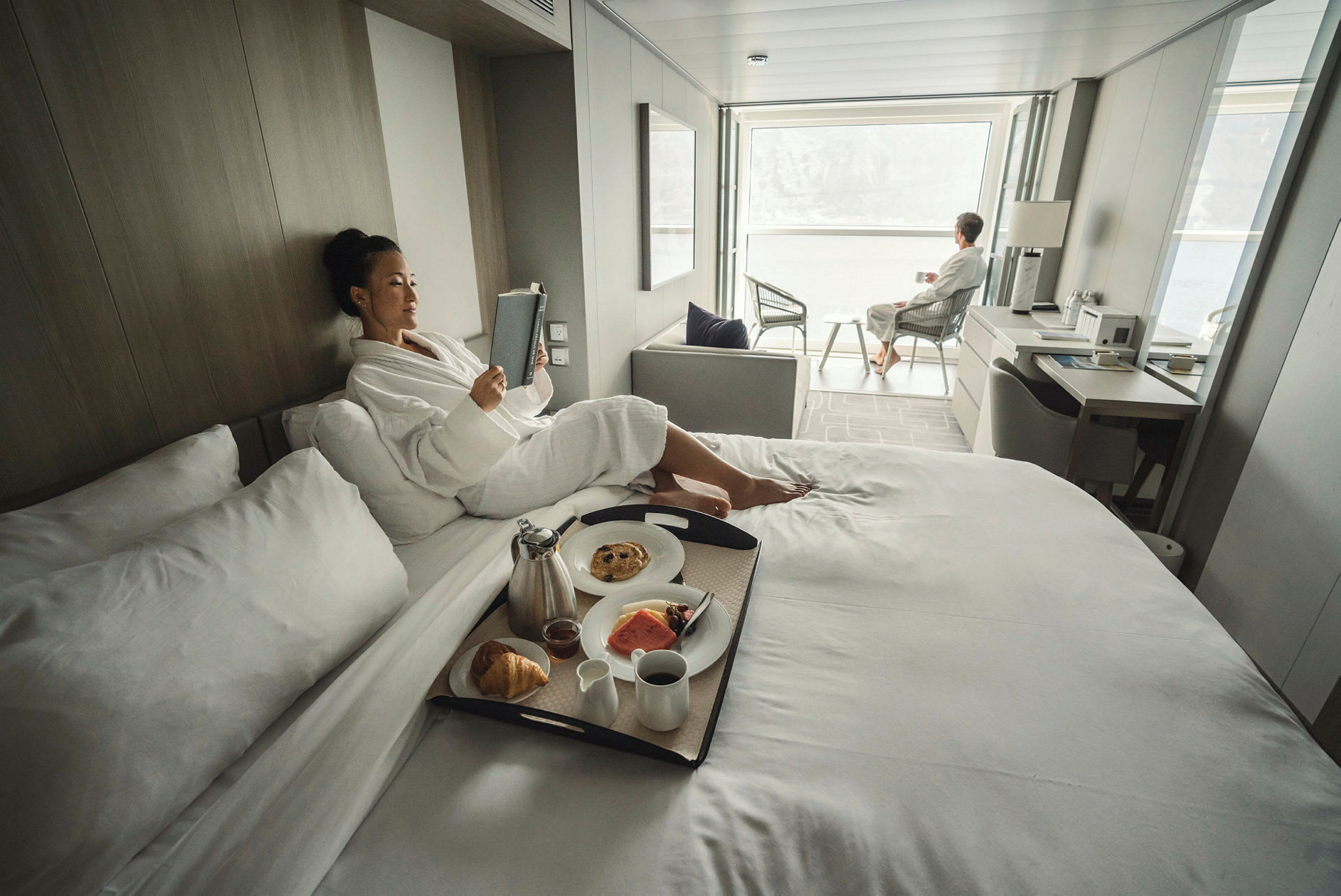 Personer slapper af i et lyst og moderne hotelværelse, med morgenmad serveret på sengen og siddeplads ved en stor vinduesudsigt. (AI-genereret tekst)