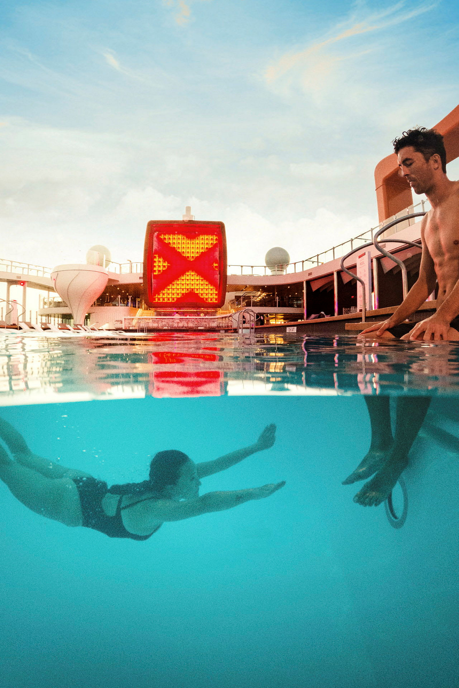 En person svømmer i en pool under vandet, mens en anden står ved poolkanten. Baggrunden viser et skibs åbne dæk og himlen. (AI-genereret tekst)