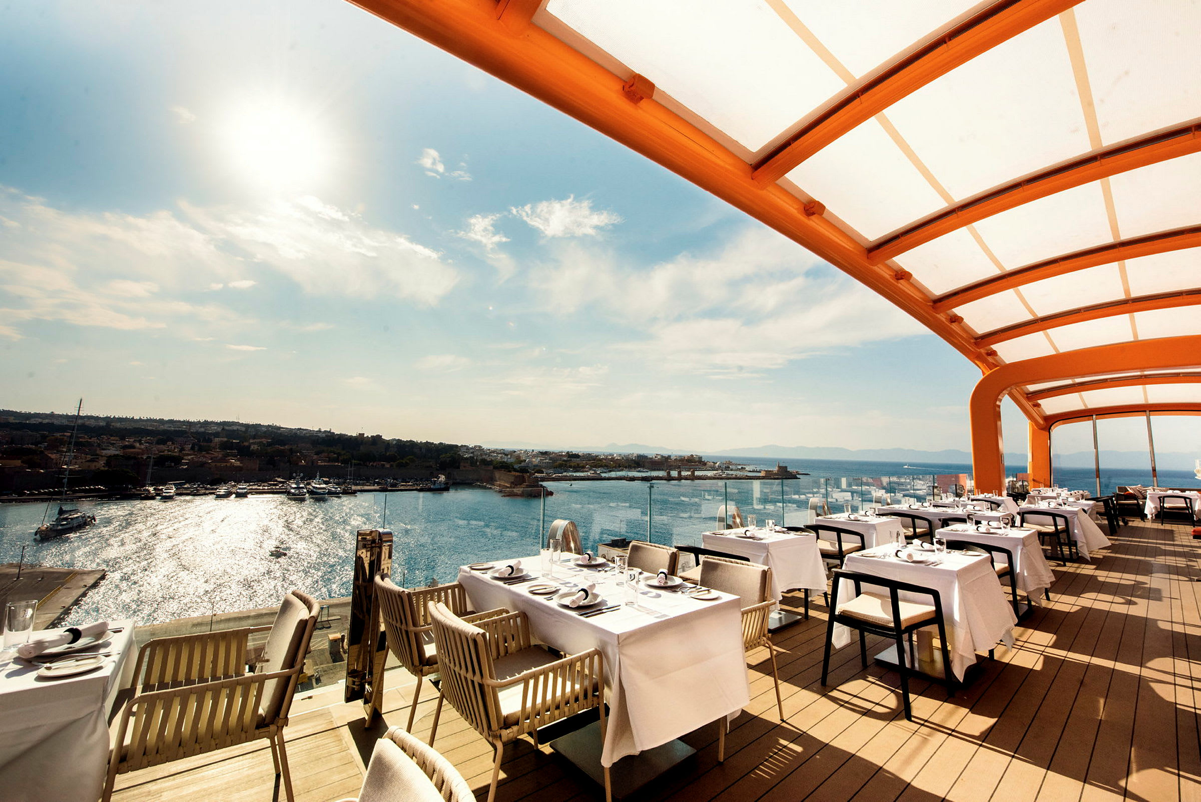 Billede af en restaurant med udendørs servering under et overdække, med havet og nogle både i baggrunden. (AI-genereret tekst)