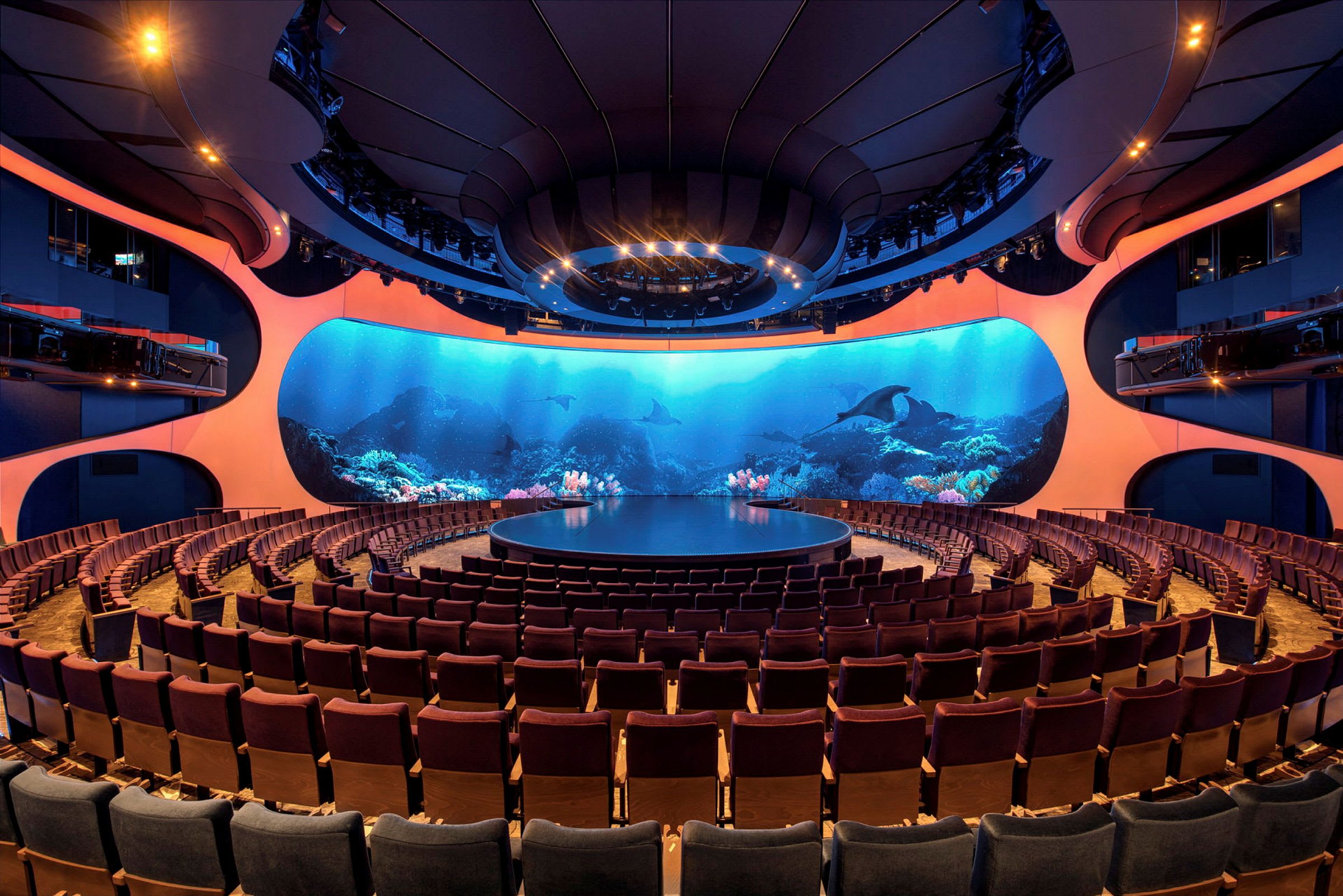 Et auditorium med en rund scene og rækker af publikumsstole. Baggrunden består af en stor skærm, der viser et undervandsmotiv. (AI-genereret tekst)