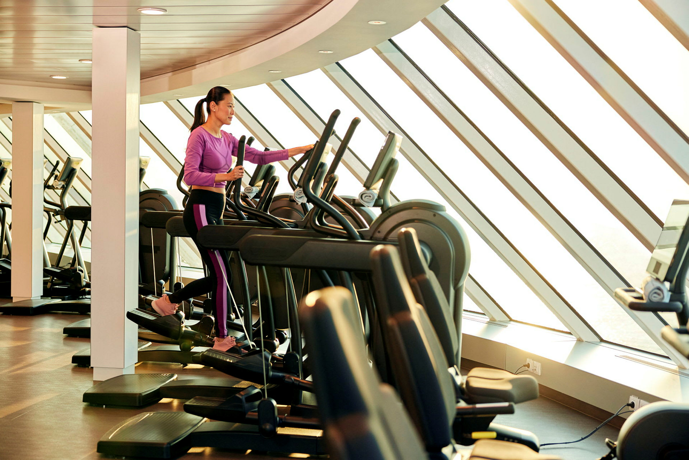 Person træner på en elliptisk træningsmaskine i et fitnesscenter med store vinduer. (AI-genereret tekst)