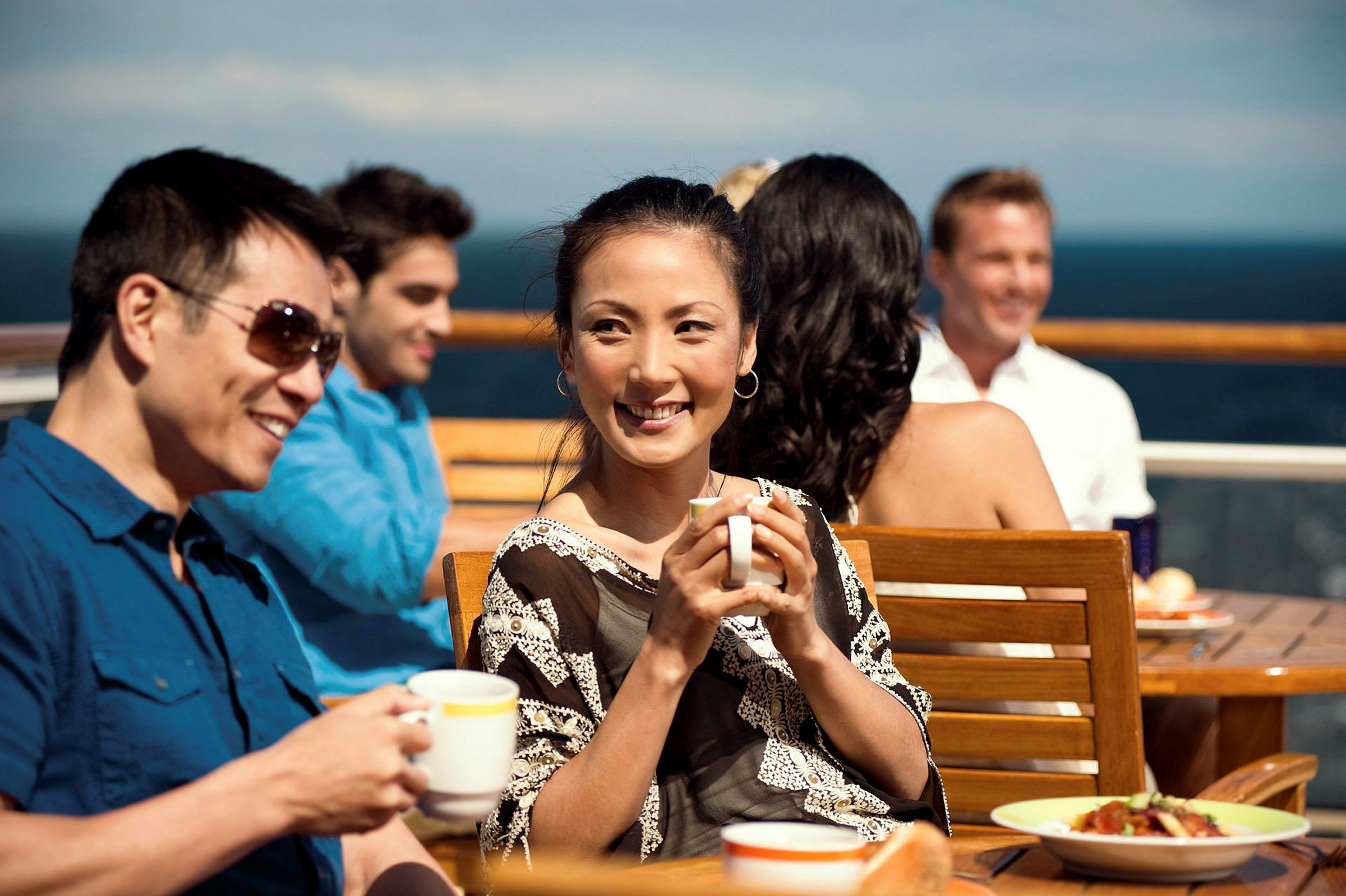 Personer sidder ved et bord og drikker varme drikke på en terrasse med udsigt til havet. (AI-genereret tekst)