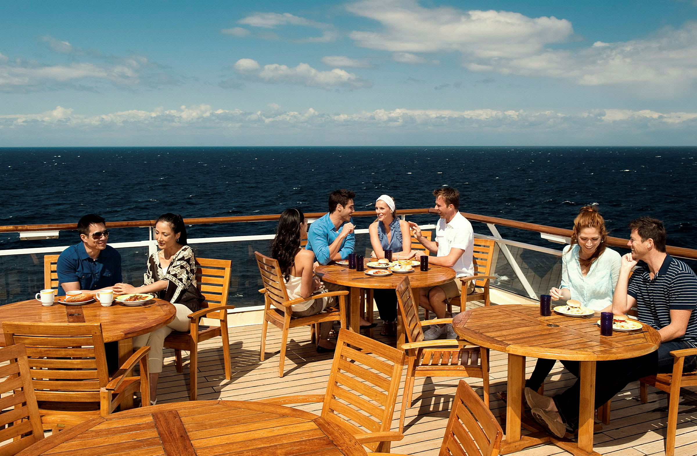 Mennesker sidder ved træmøbler på en udendørs terrasse med udsigt over havet. (AI-genereret tekst)
