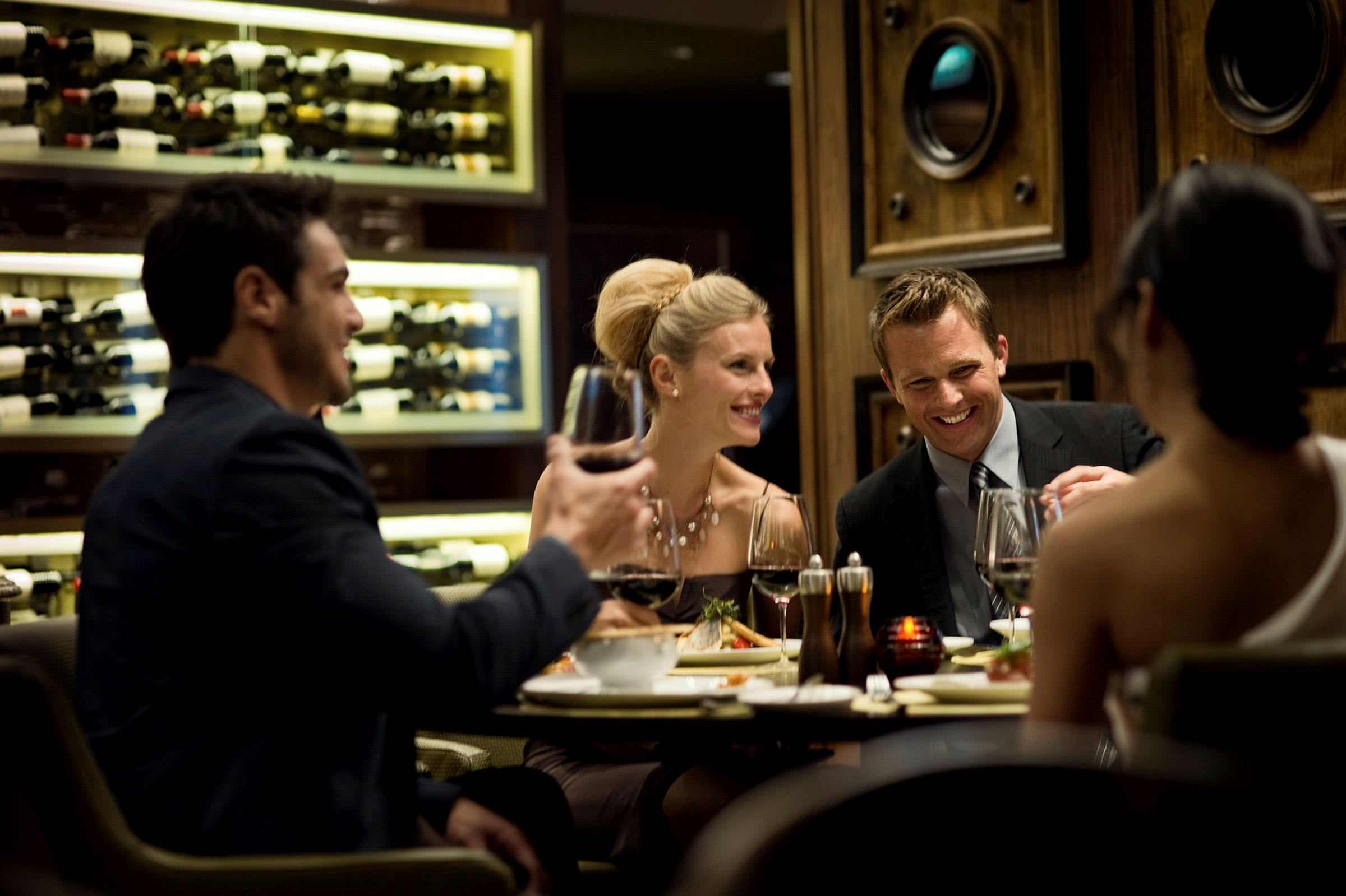 En gruppe mennesker nyder mad og vin ved et bord i en elegant restaurant med en væg dekoreret med vinforsyninger i baggrunden. (AI-genereret tekst)