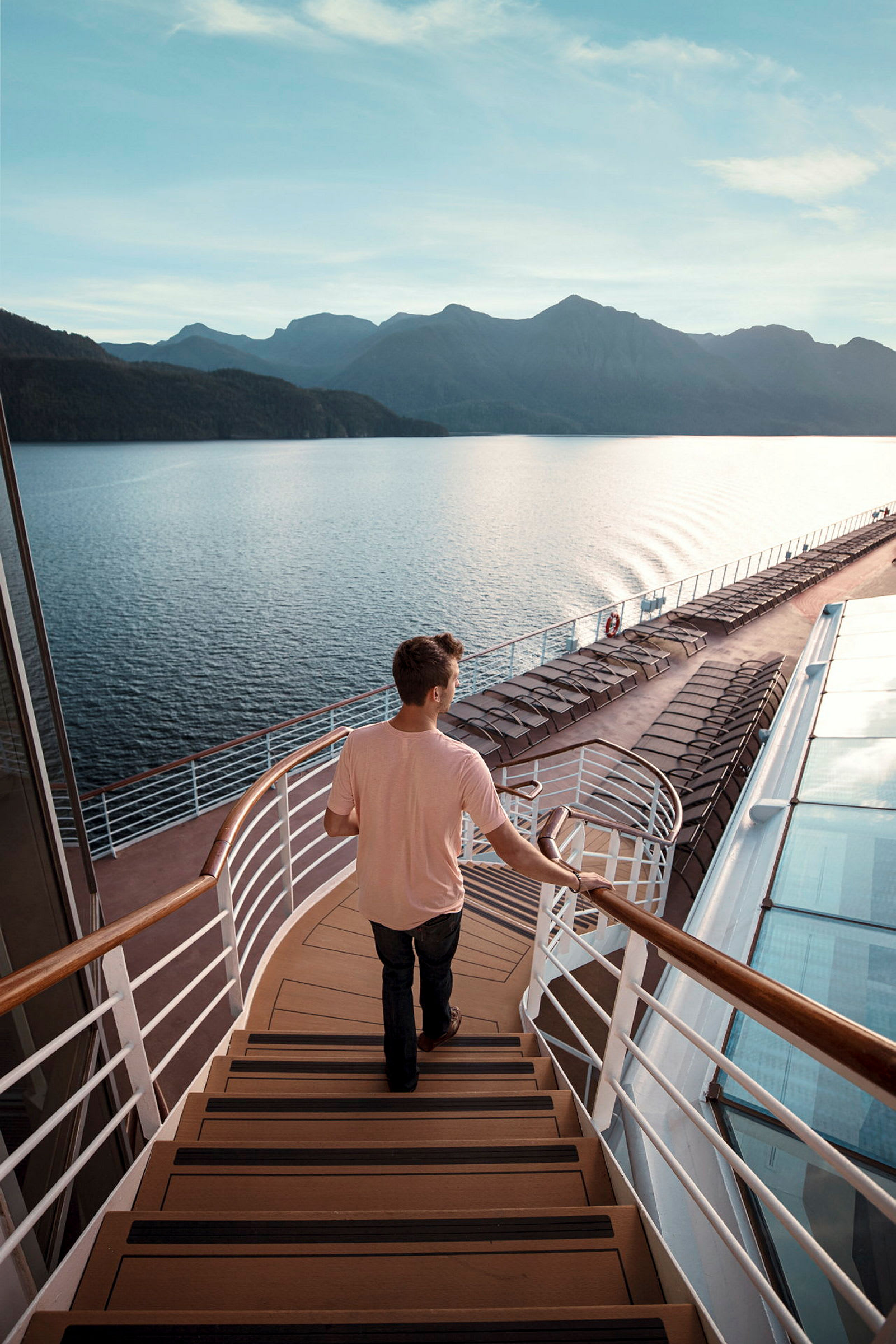 En person går ned ad en trappe på et krydstogtskib med udsigt over havet og bjergene i baggrunden. (AI-genereret tekst)