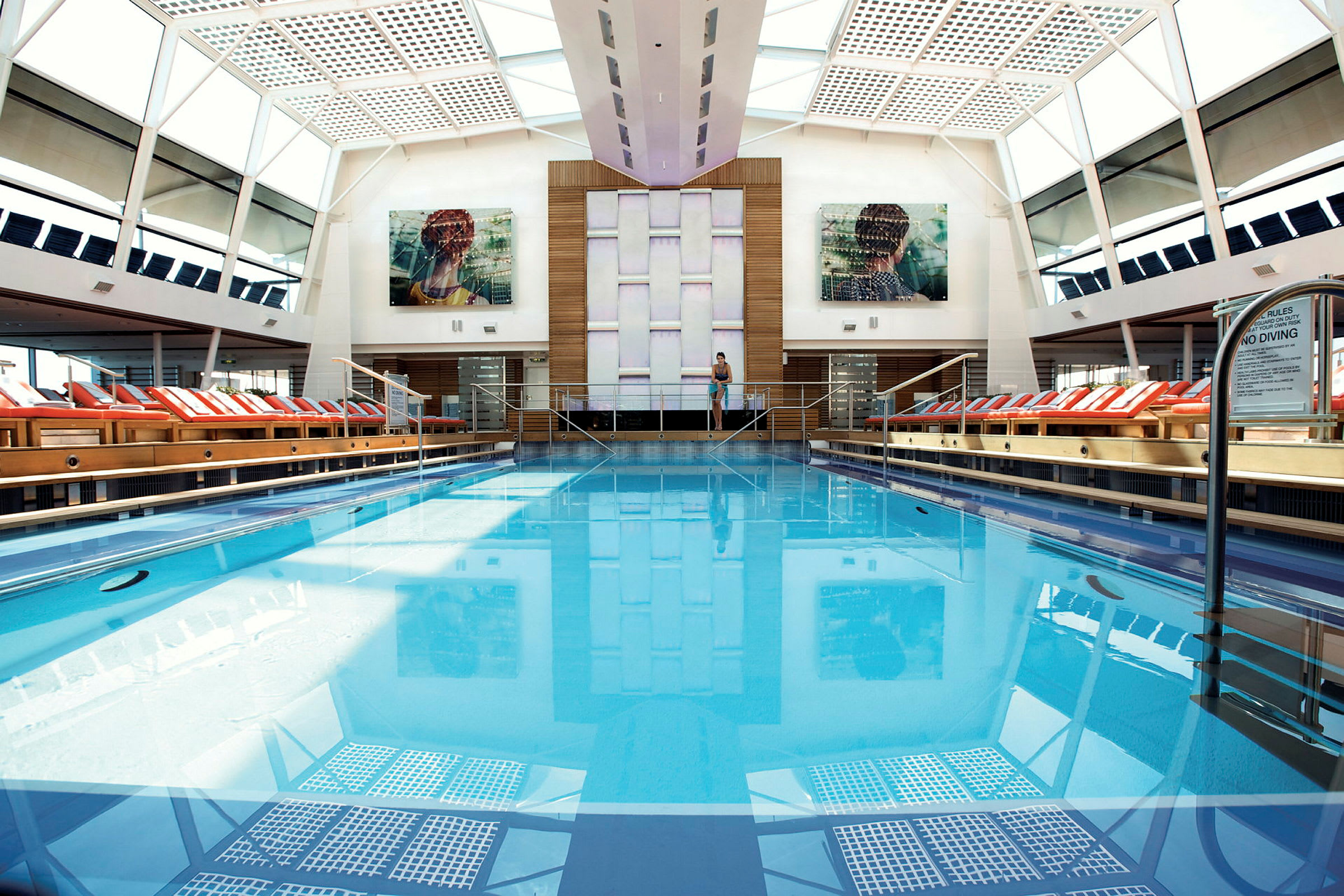 Indendørs pool med klart blåt vand omgivet af liggestole under et glastag. (AI-genereret tekst)