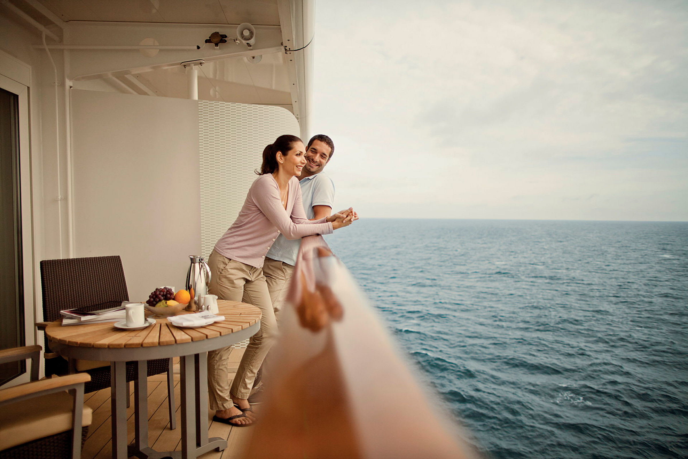 Personer står på en balkon med udsigt over havet. Et træbord er placeret med frugt og drikke. (AI-genereret tekst)