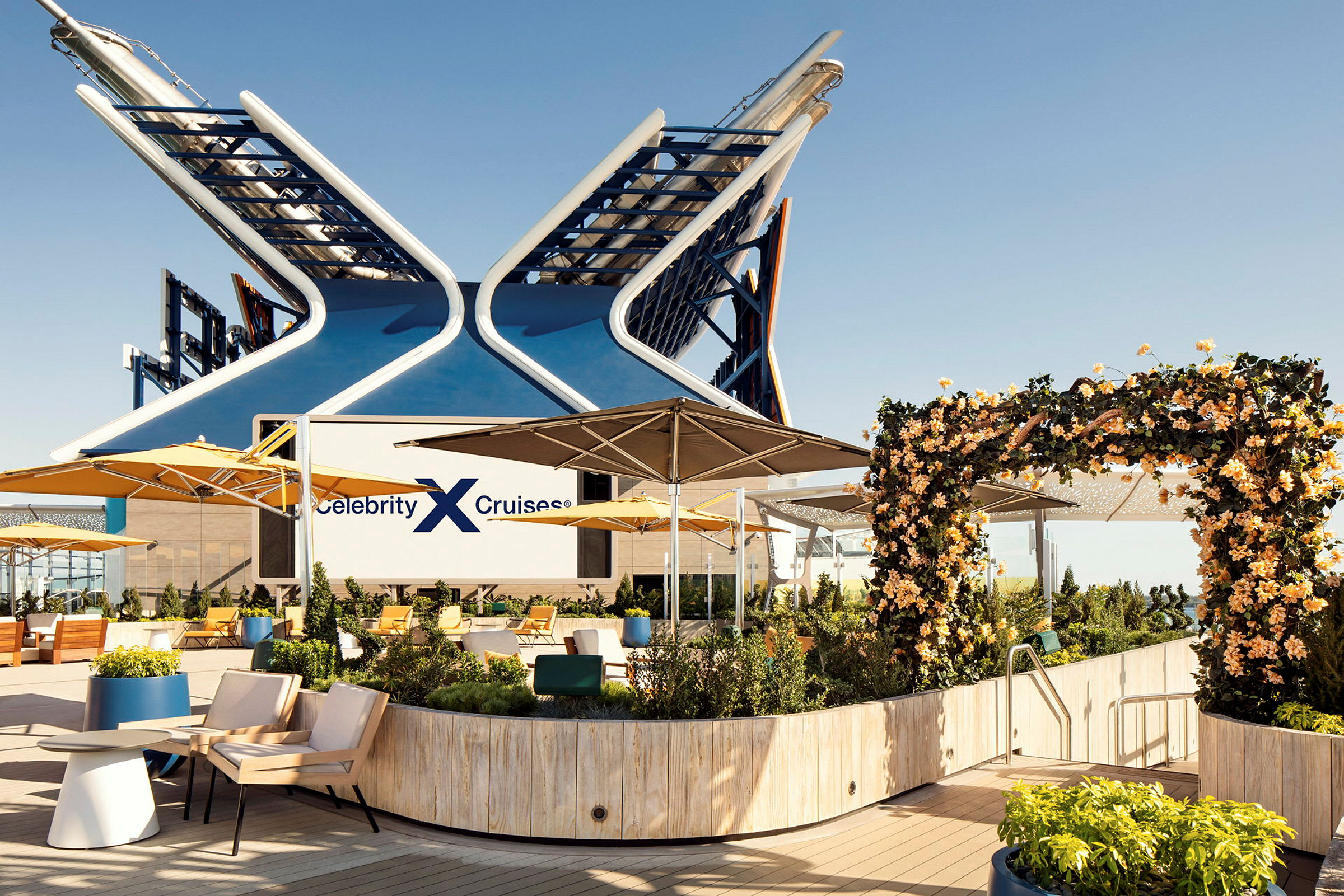 Et skibsdæk med siddeområder, gule parasoller og velplejet beplantning, med logoet 'Celebrity Cruises' synligt. (AI-genereret tekst)