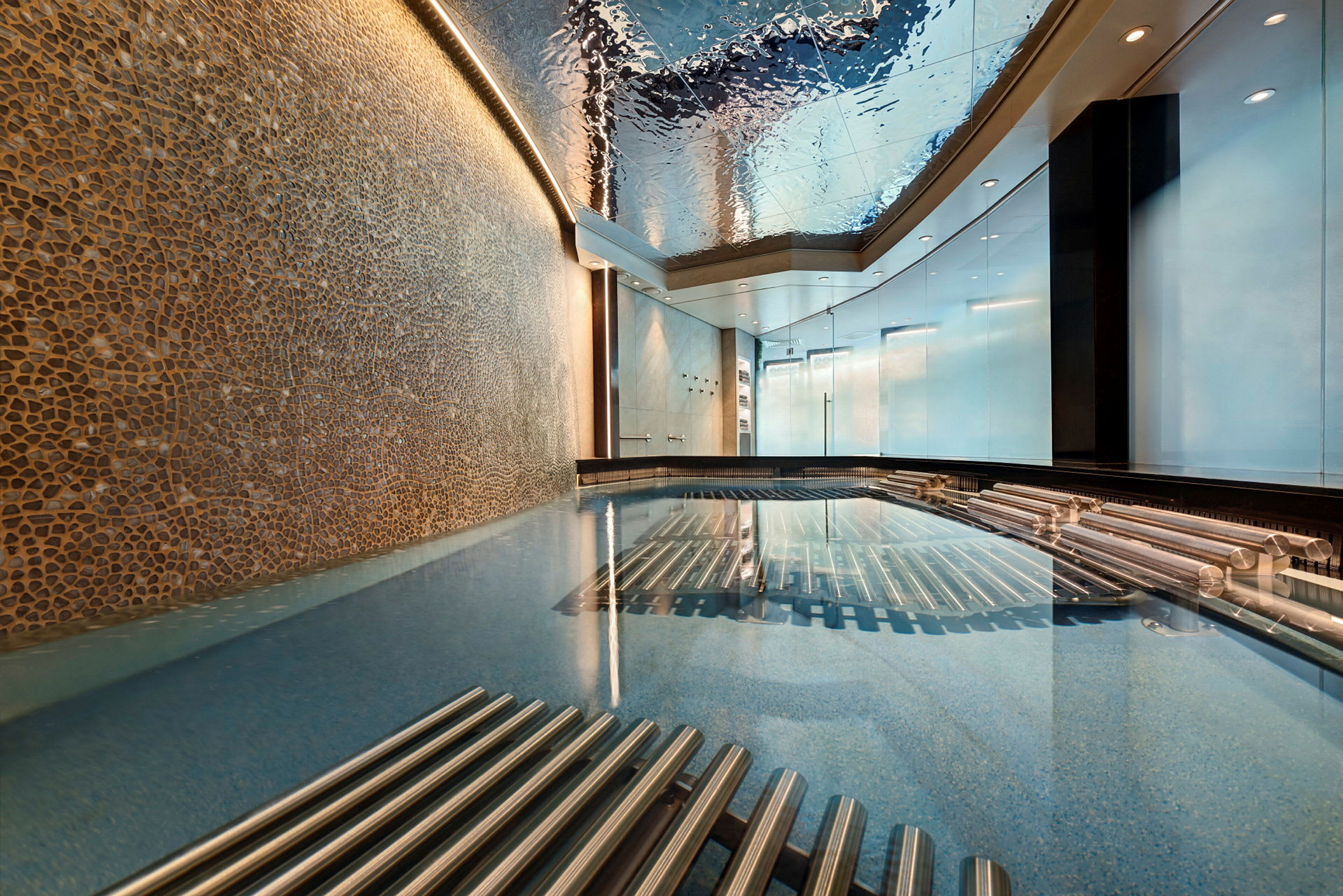 En indendørs pool med et loft, der reflekterer lys og omgivelser, flankeret af metalliske detaljer og vægge med dekorative mosaikker. (AI-genereret tekst)
