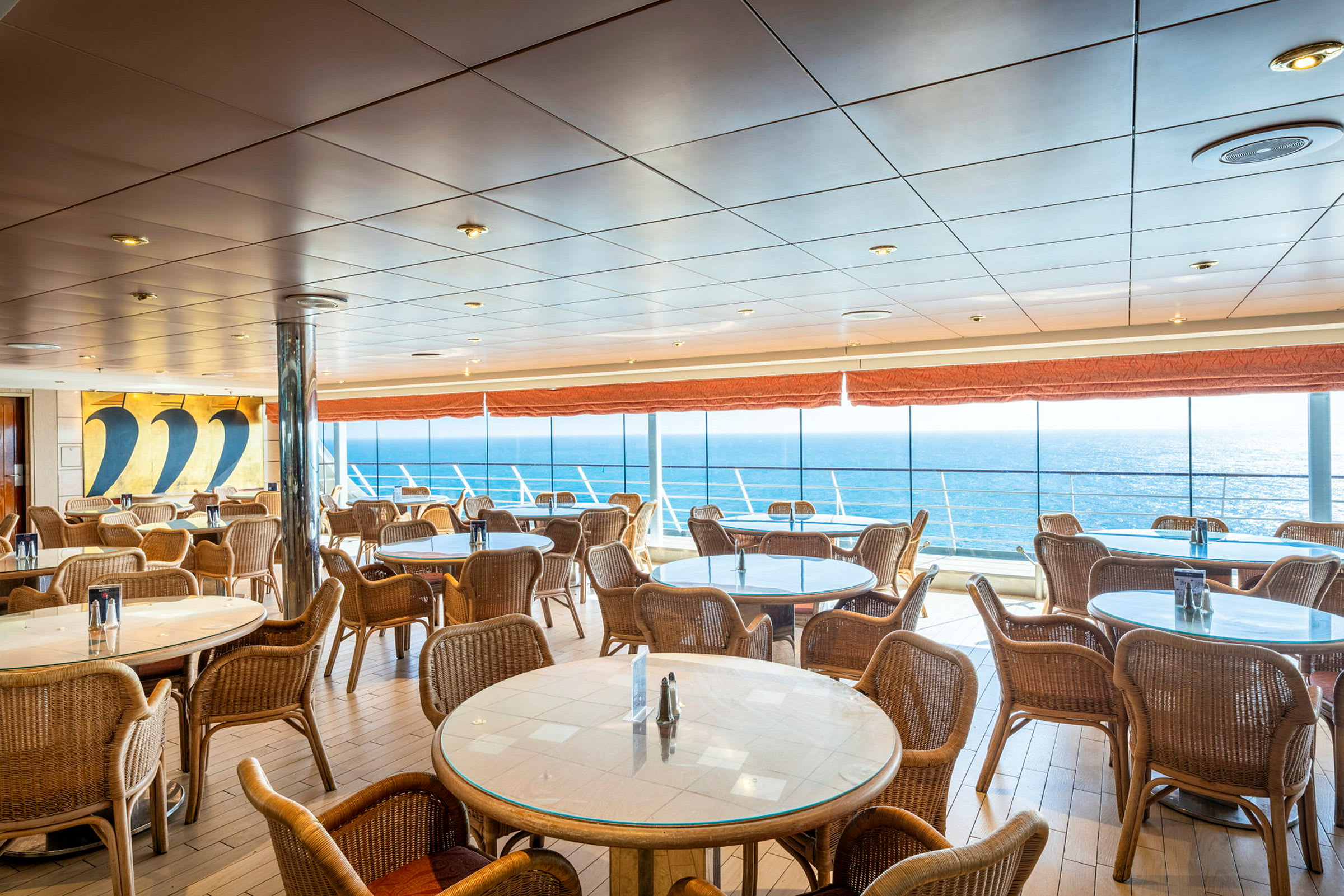 Spiseområde med runde borde og stole samt store vinduer med udsigt over havet. (AI-genereret tekst)