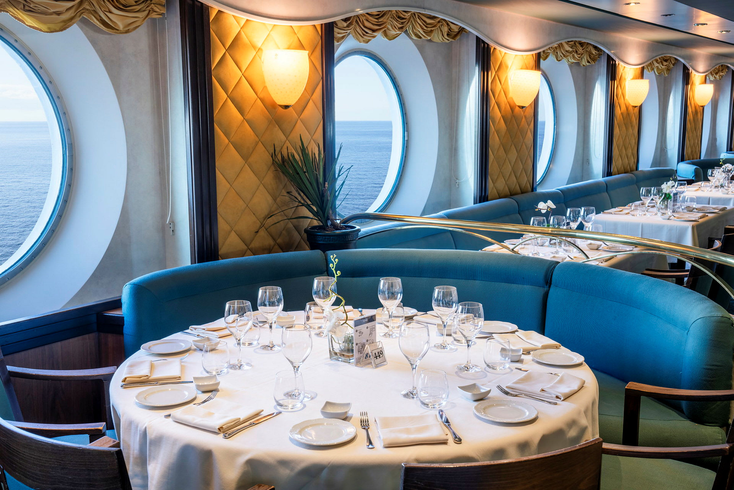En restaurant med dækkede borde, blå sofaer og store runde vinduer, der viser udsigt over havet. (AI-genereret tekst)