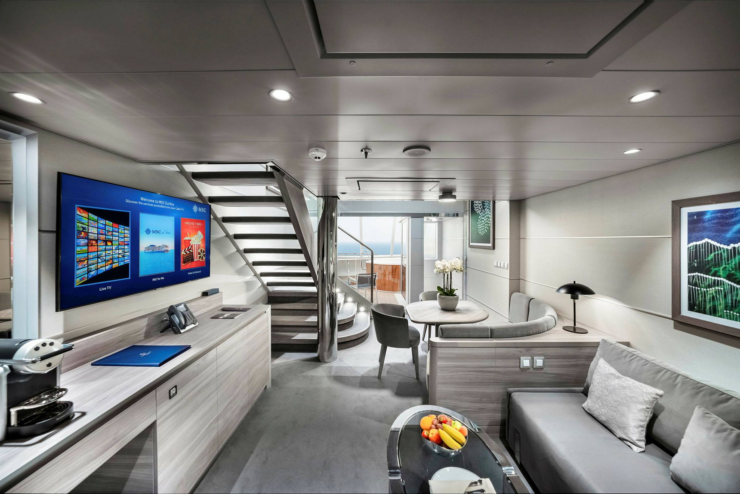 En moderne og lys indretning i en hotelsuite med sofa, bord, fladskærms-tv og trappe, der fører til en øvre etage samt en balkon med udsigt. (AI-genereret tekst)