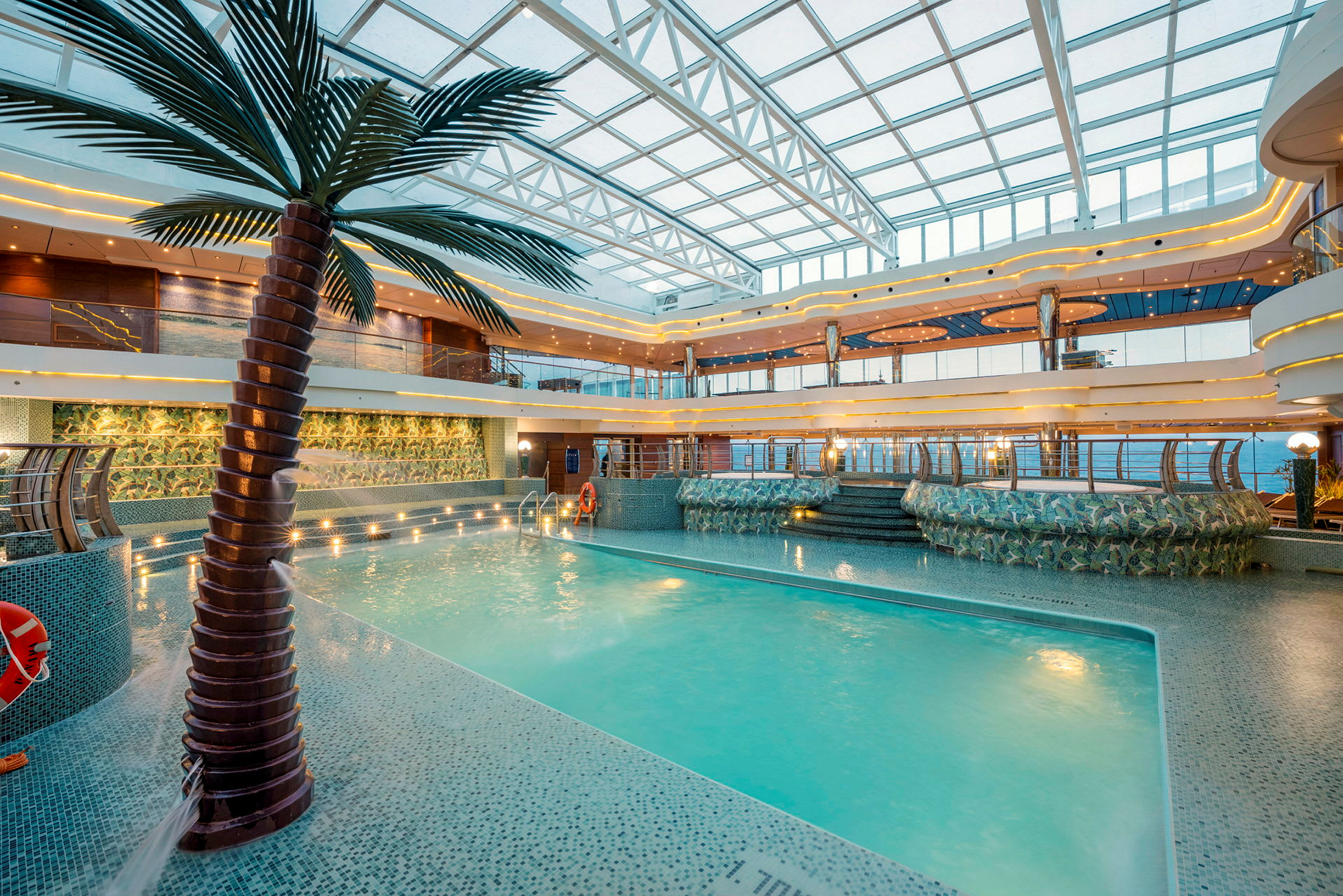 En indendørs pool omgivet af tropisk dekorativ design, med siddepladser og trapper omkring. (AI-genereret tekst)