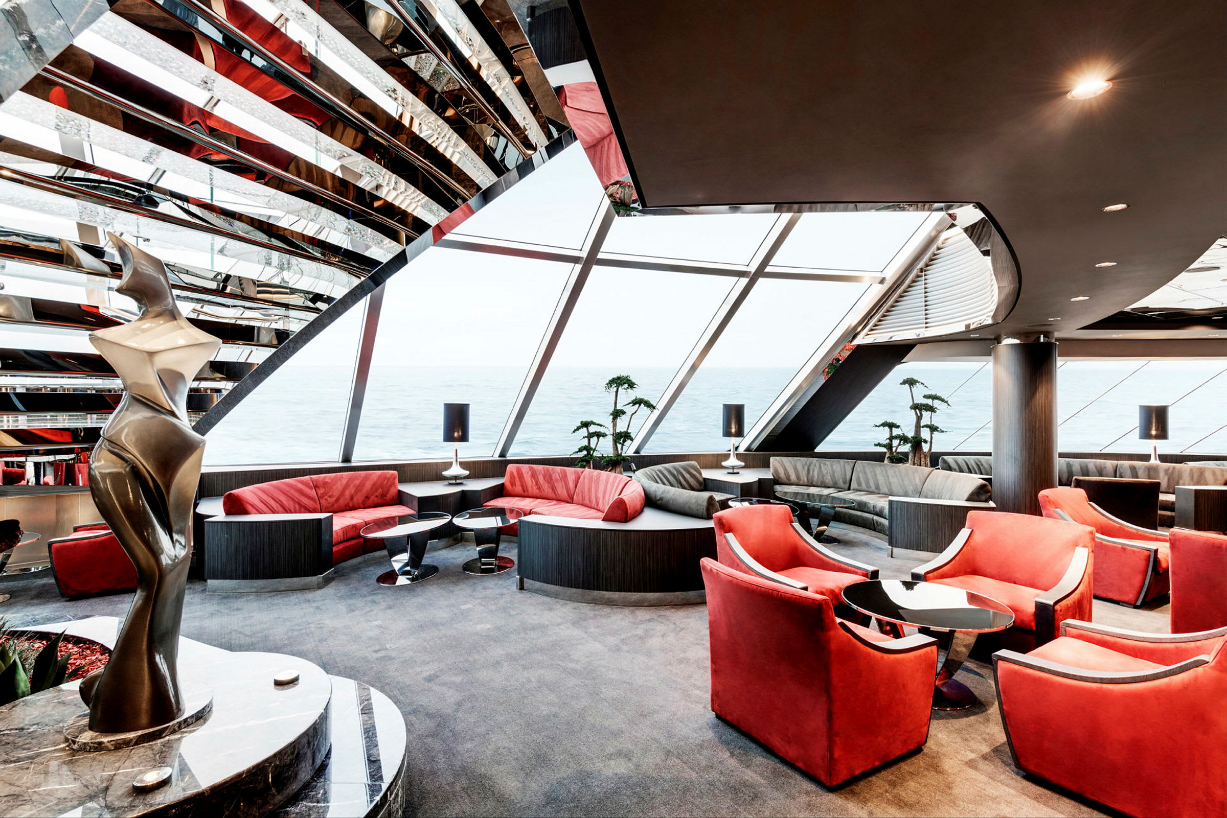 Et moderne indrettet rum med røde stole og sofaer, store vinduer med udsigt over havet samt en abstrakt skulptur ved siden. (AI-genereret tekst)