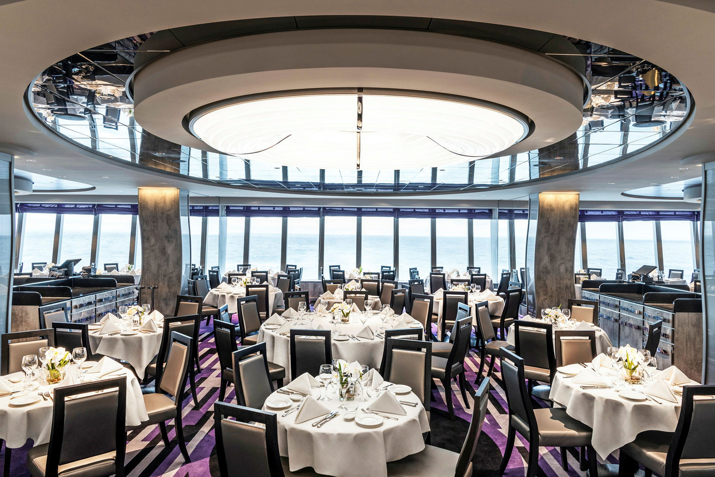 Billede af en elegant restaurant med borde dækket med hvide duge og stole, vinduer med havudsigt ses i baggrunden. (AI-genereret tekst)