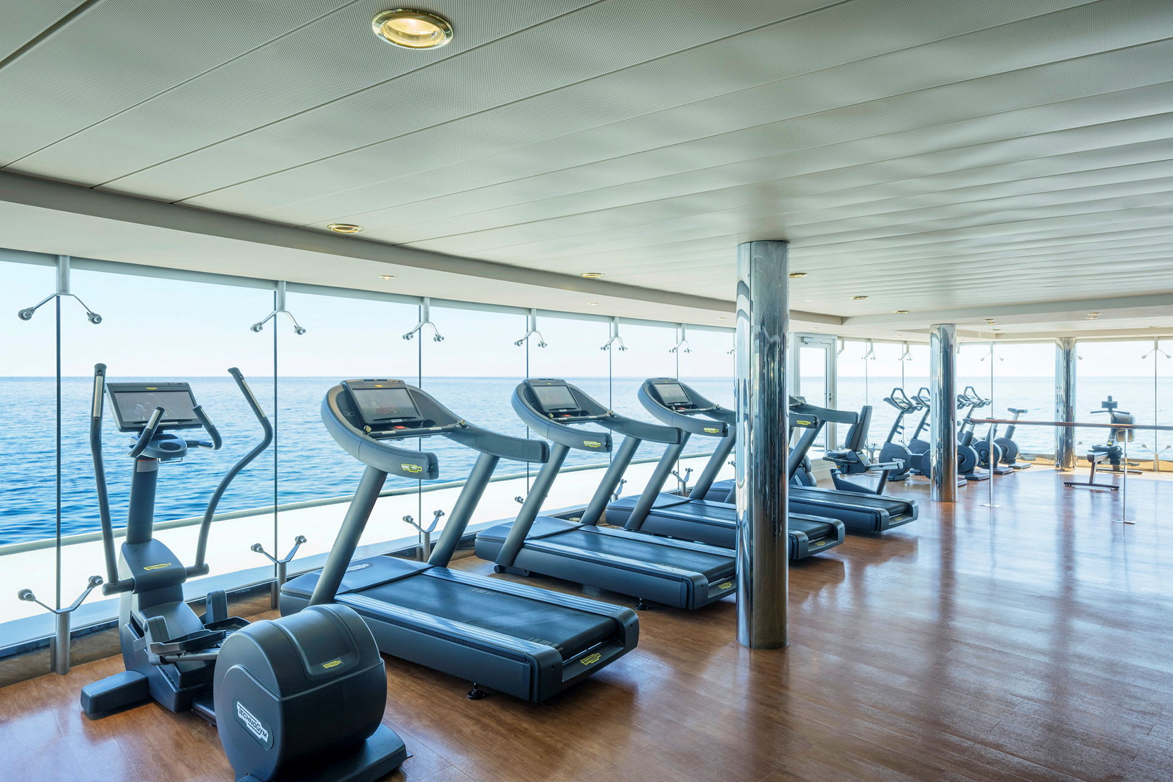 Billede af et fitnesscenter med træningsmaskiner ved store vinduer med udsigt over havet. (AI-genereret tekst)