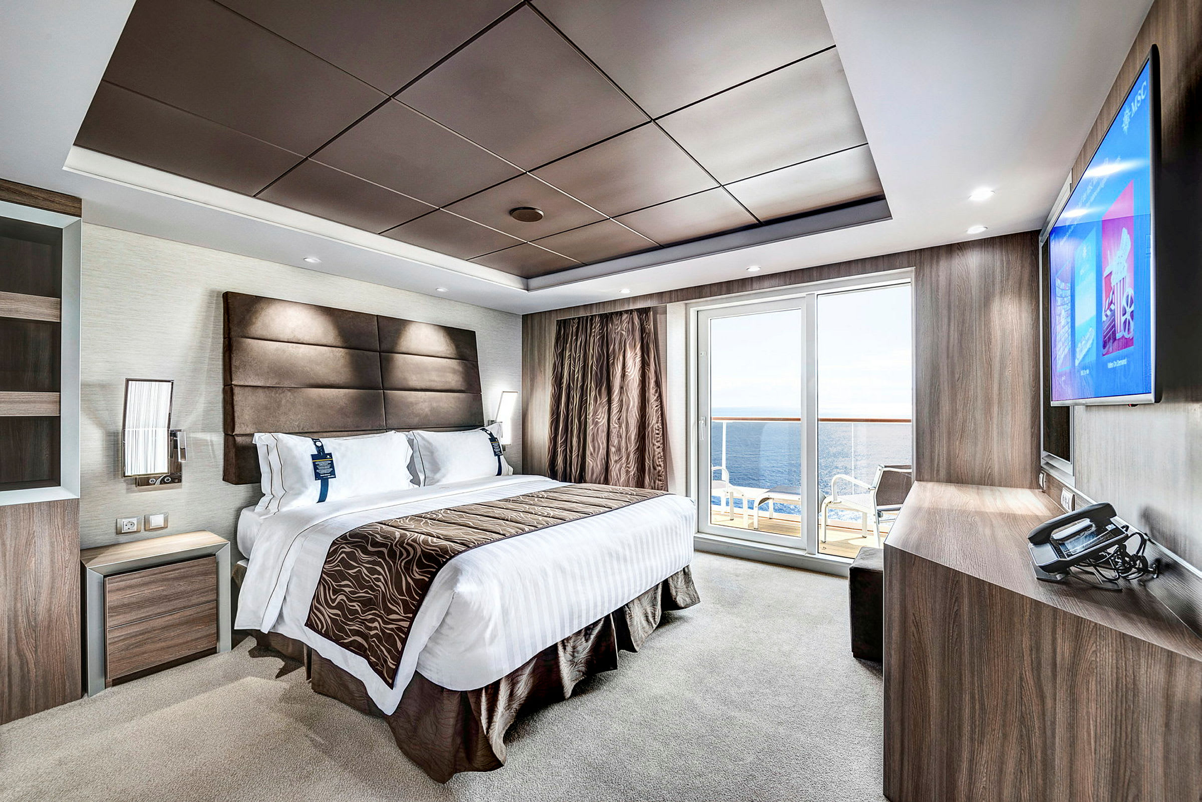 Et moderne hotelværelse med en stor seng, en fladskærms-TV, et skrivebord og en altan med udsigt til havet. (AI-genereret tekst)