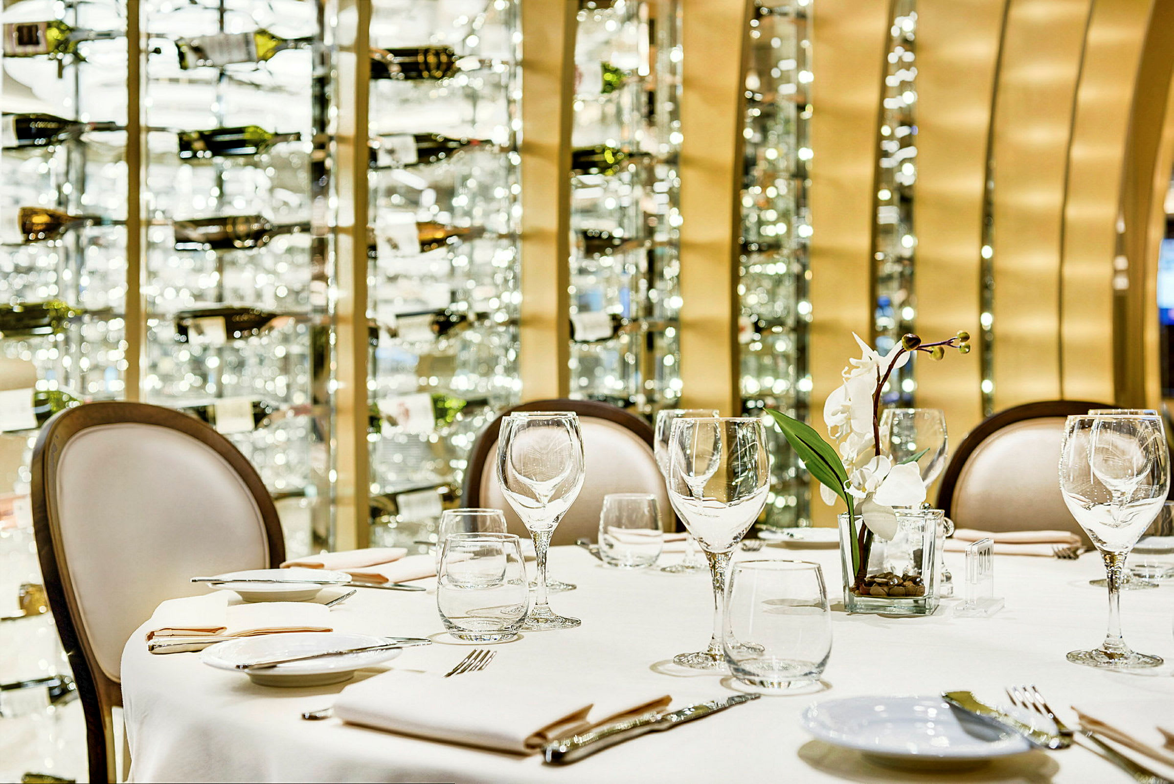 Borddækning med glas, tallerkener og en hvid blomst på en restaurant med vinflasker i baggrunden. (AI-genereret tekst)