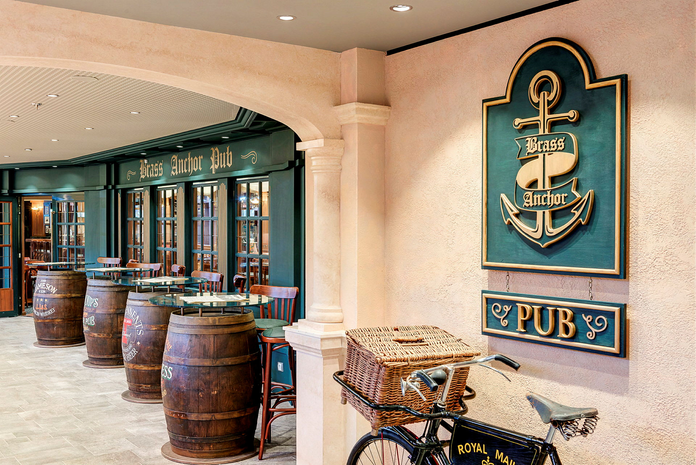 Interiør af en pub med et skilt, der siger 'Brass Anchor Pub', træmøbler og tønder, der bruges som borde, samt en cykel med en kurv foran. (AI-genereret tekst)