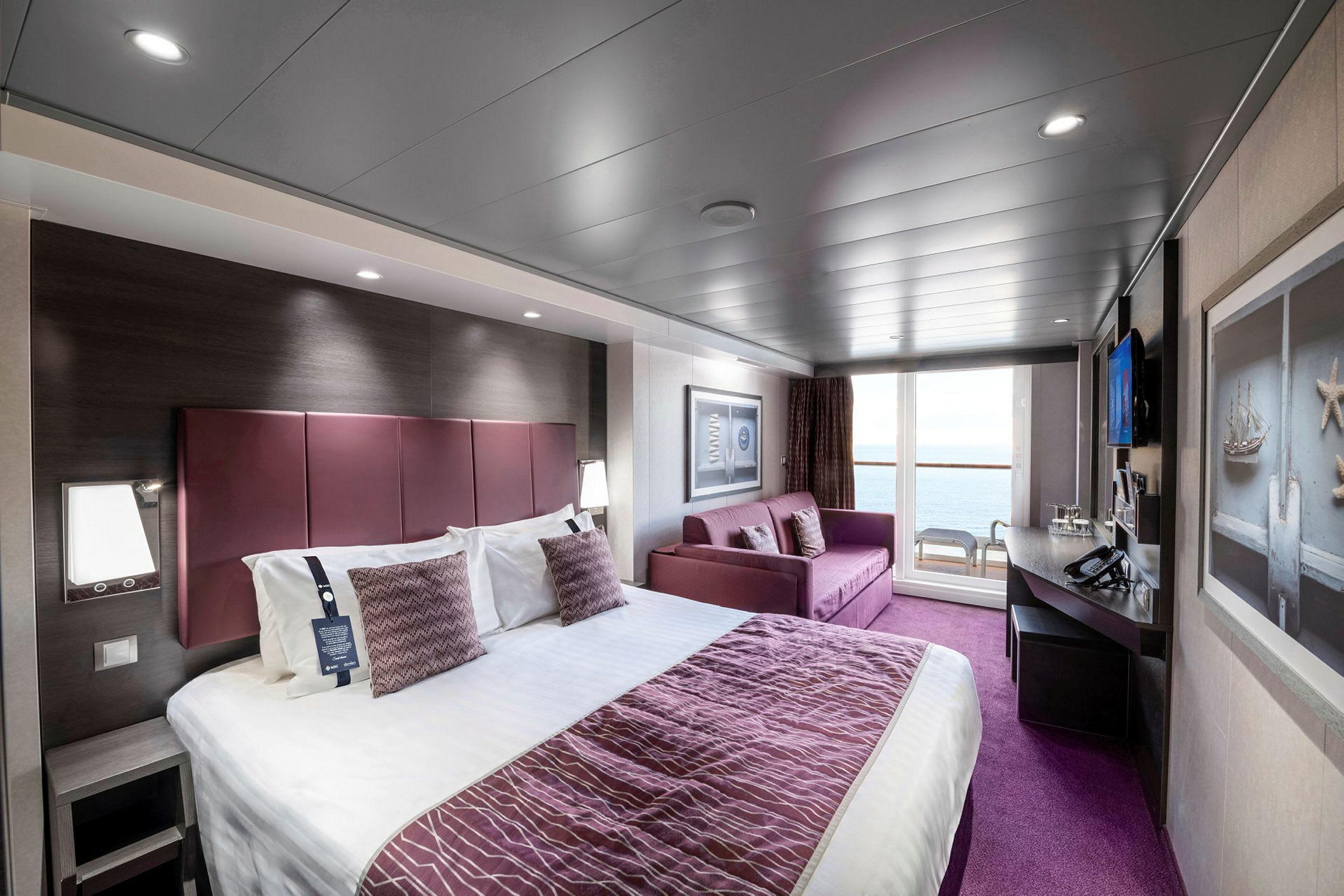 Et hotelværelse med en stor seng, en sofa, skrivebord og udsigt gennem glasdøre mod en balkon med stole. (AI-genereret tekst)