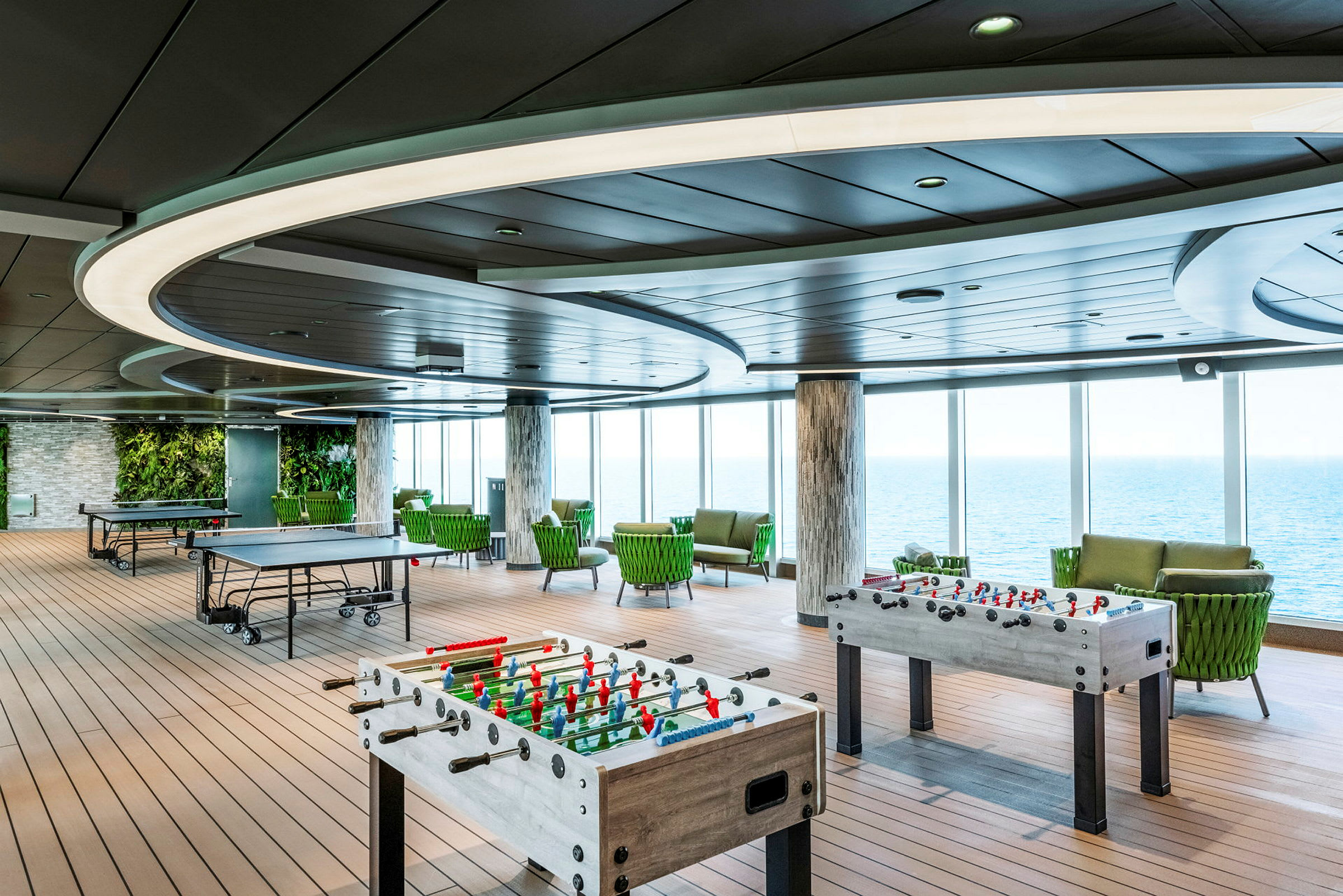 Et rummeligt rum med bordtennisborde og bordfodbold, omgivet af store vinduer med udsigt over havet. (AI-genereret tekst)