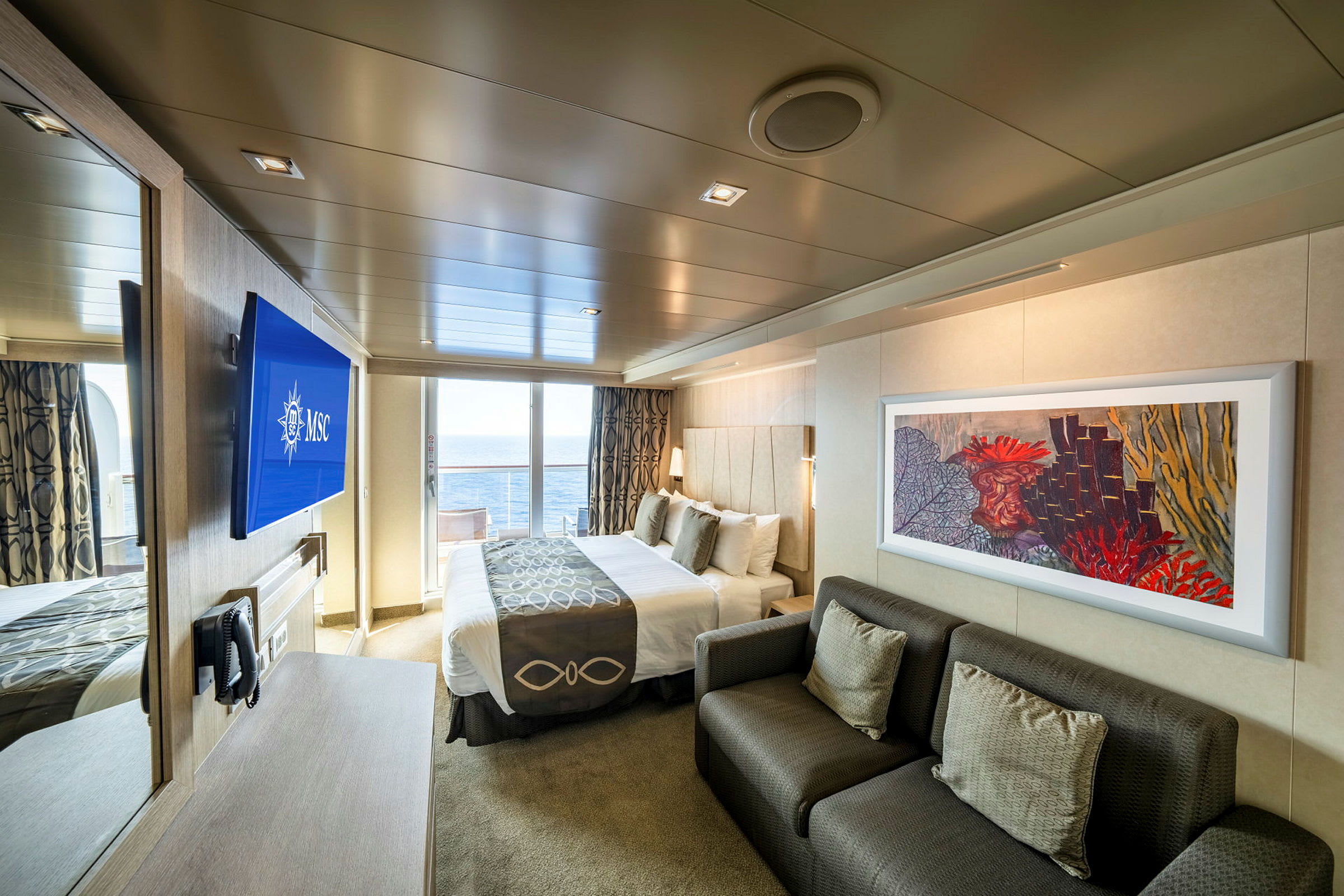 En moderne hotelsuite med havudsigt gennem glasdøre, inklusive en dobbeltseng, en sofa og en væg-tv. (AI-genereret tekst)