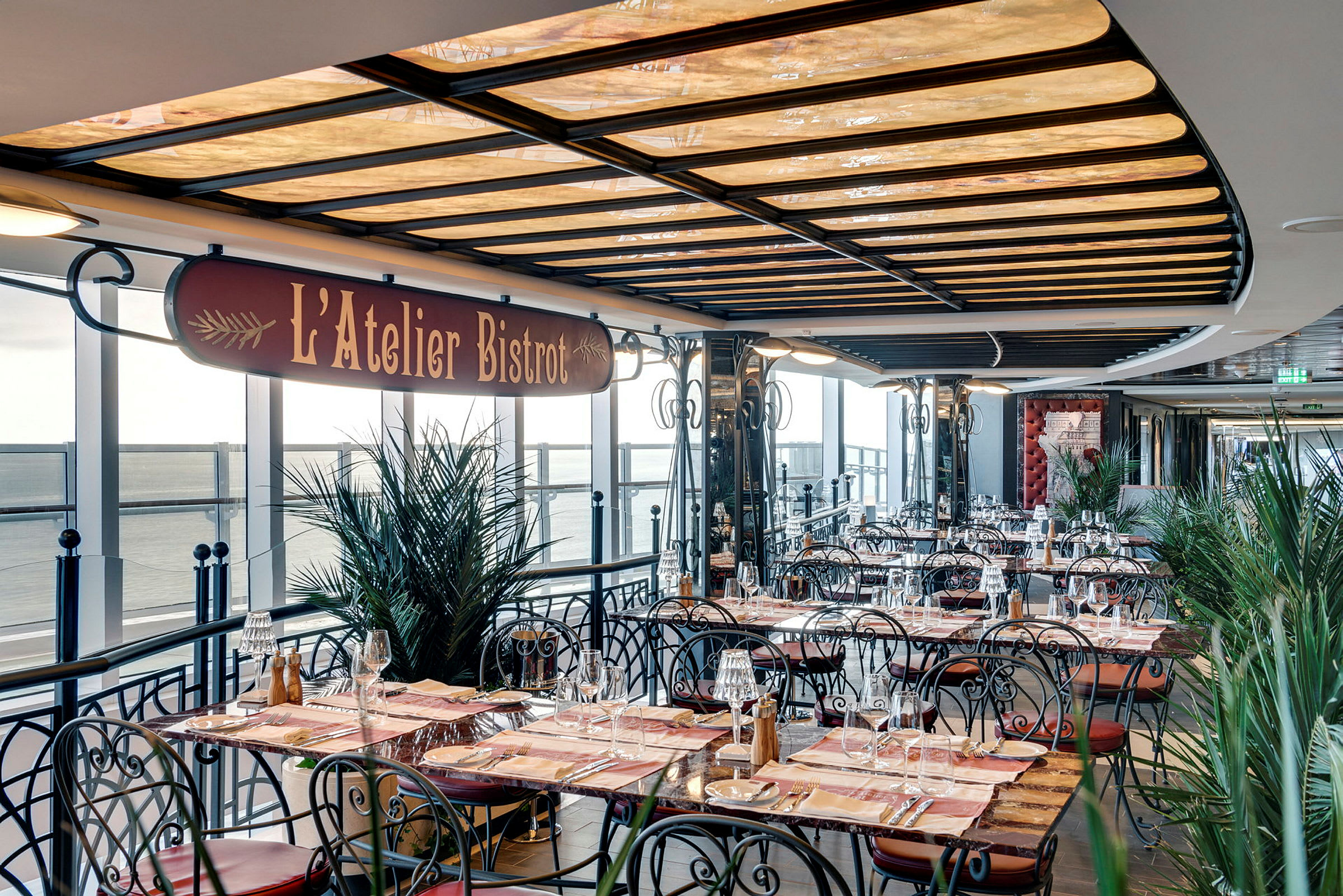Udendørs serveringsområde til restauranten L'Atelier Bistro med borde og stole og udsigt over havet. (AI-genereret tekst)