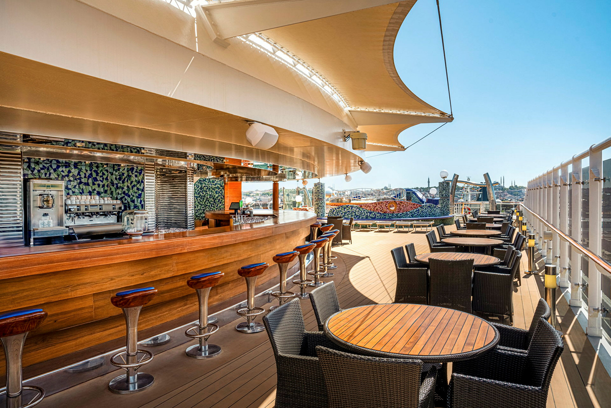 Billede af en udendørs bar med træinteriør og siddepladser omgivet af borde og stole, under en stor sejldug med udsigt over byens skyline. (AI-genereret tekst)