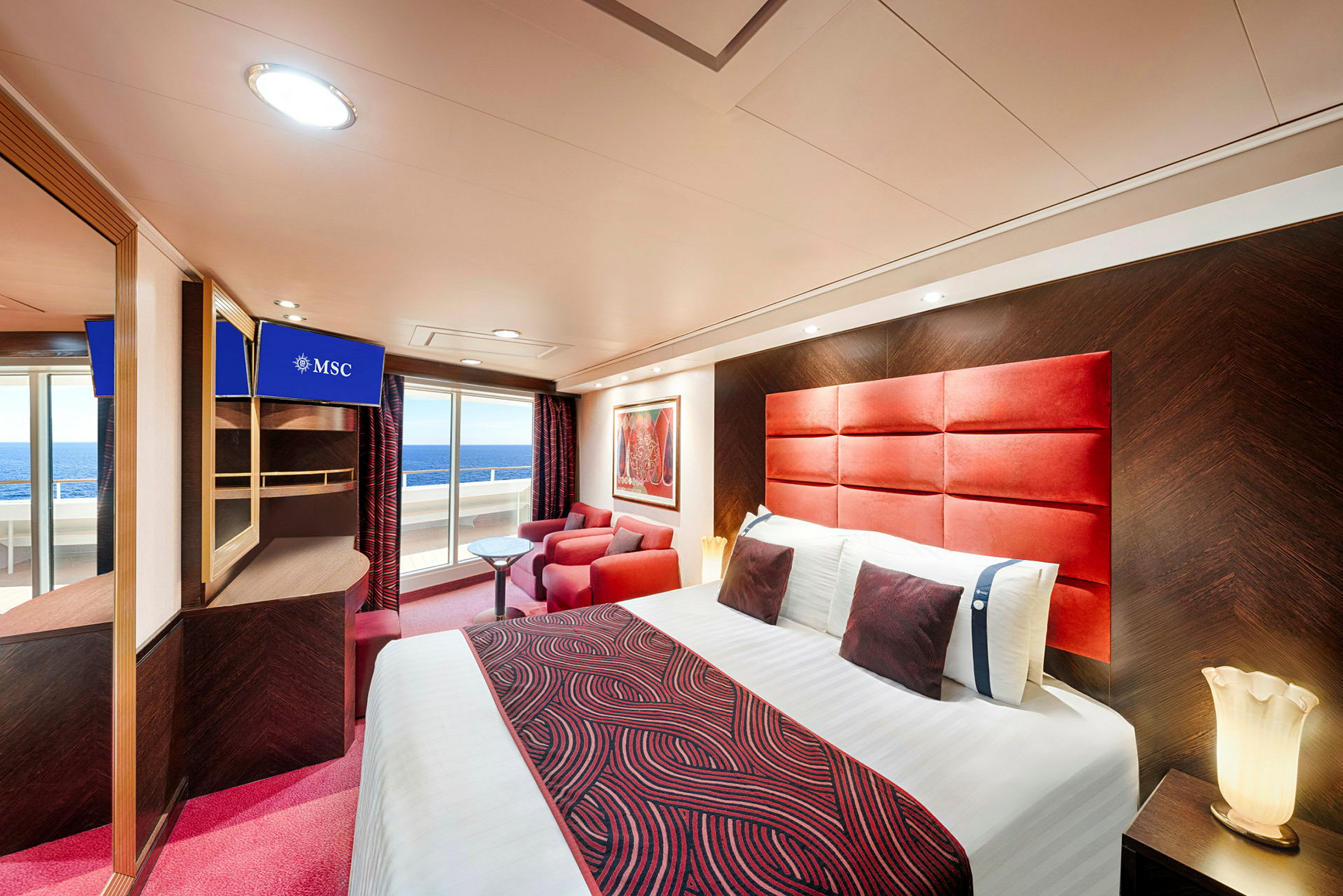 Et moderne hotelværelse med en dobbeltseng, rødmønstrede tekstiler, røde lænestole, et lille bord og en balkon med udsigt over havet. (AI-genereret tekst)