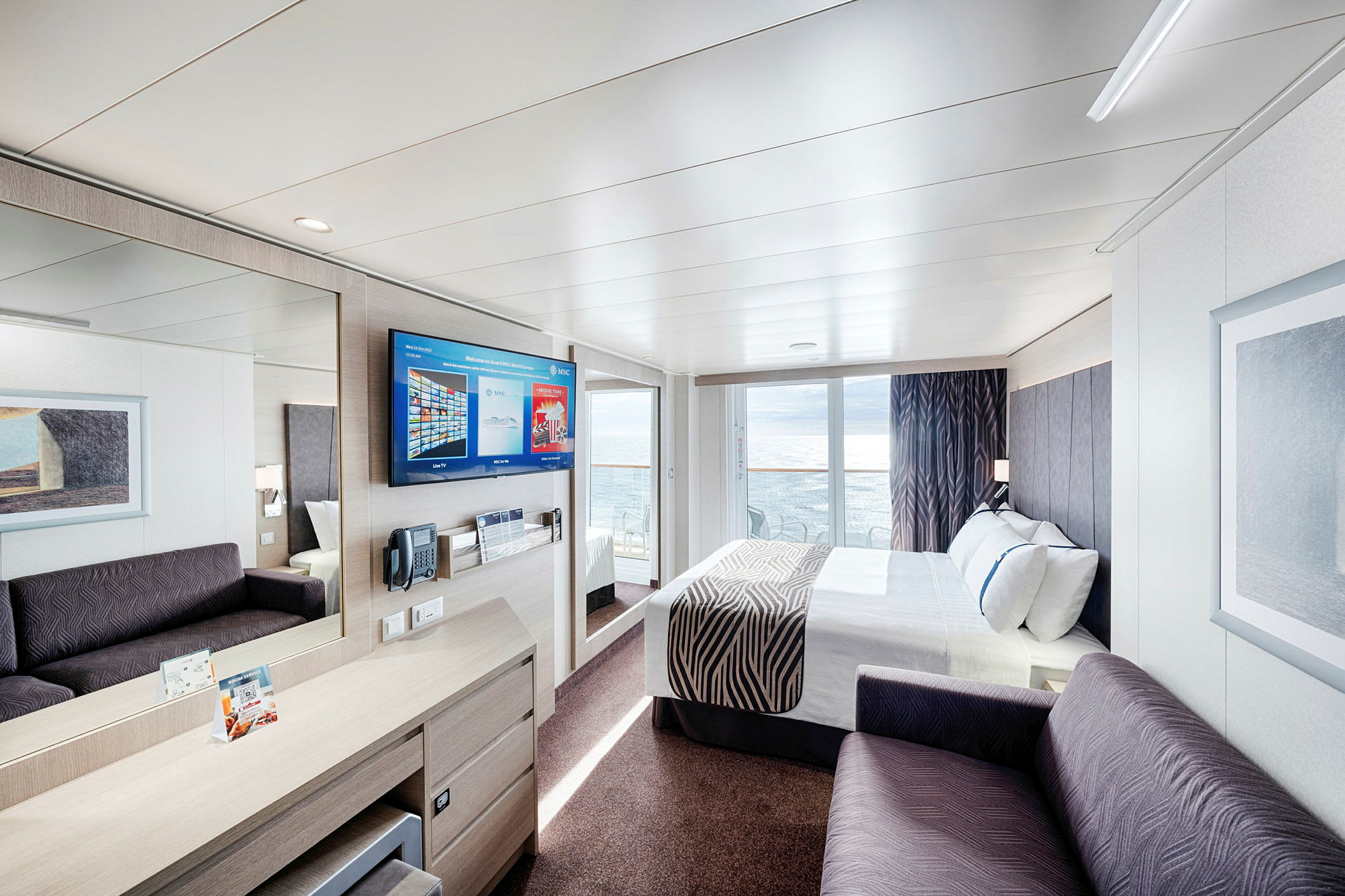 Et moderne hotelværelse med seng, sofa og et tv på væggen. Glasdøre fører ud til en balkon med havudsigt. (AI-genereret tekst)