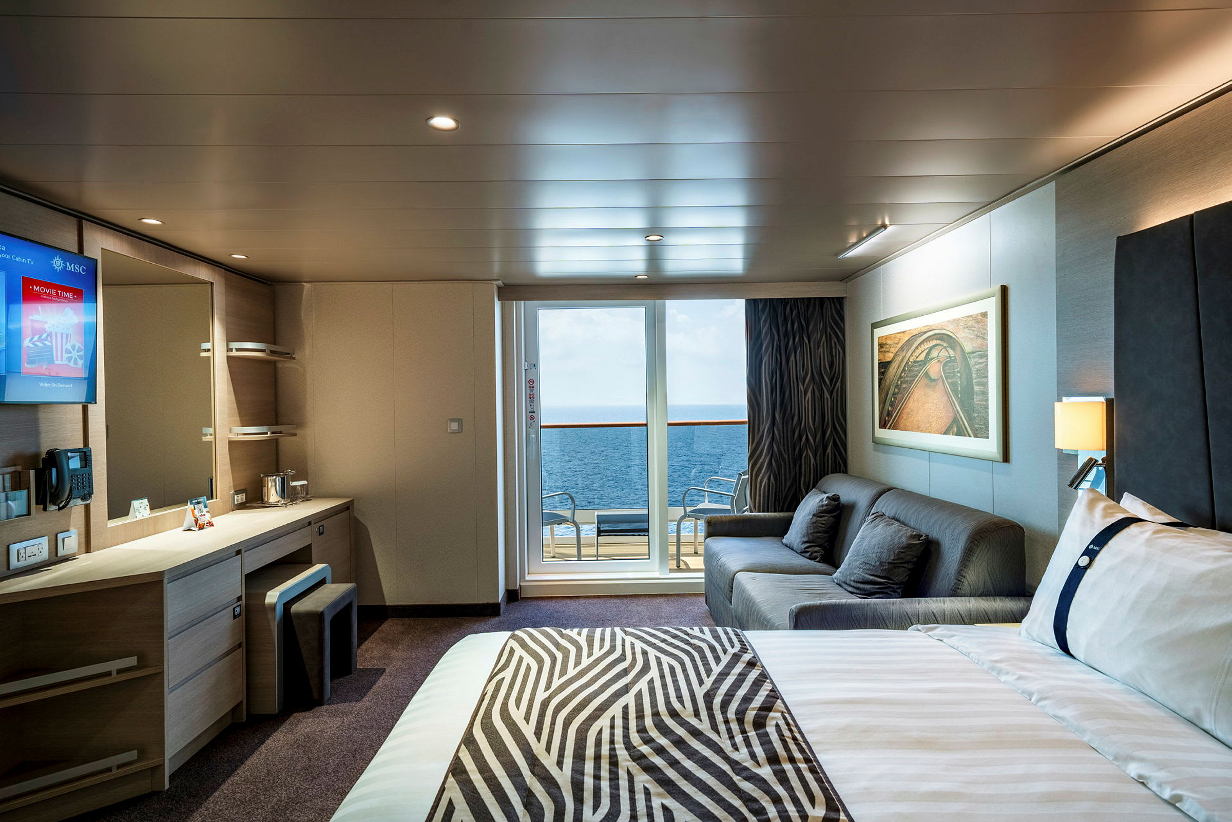 En interiør af et hotelværelse med seng, sofa, skrivebord og en altan med udsigt over havet. (AI-genereret tekst)
