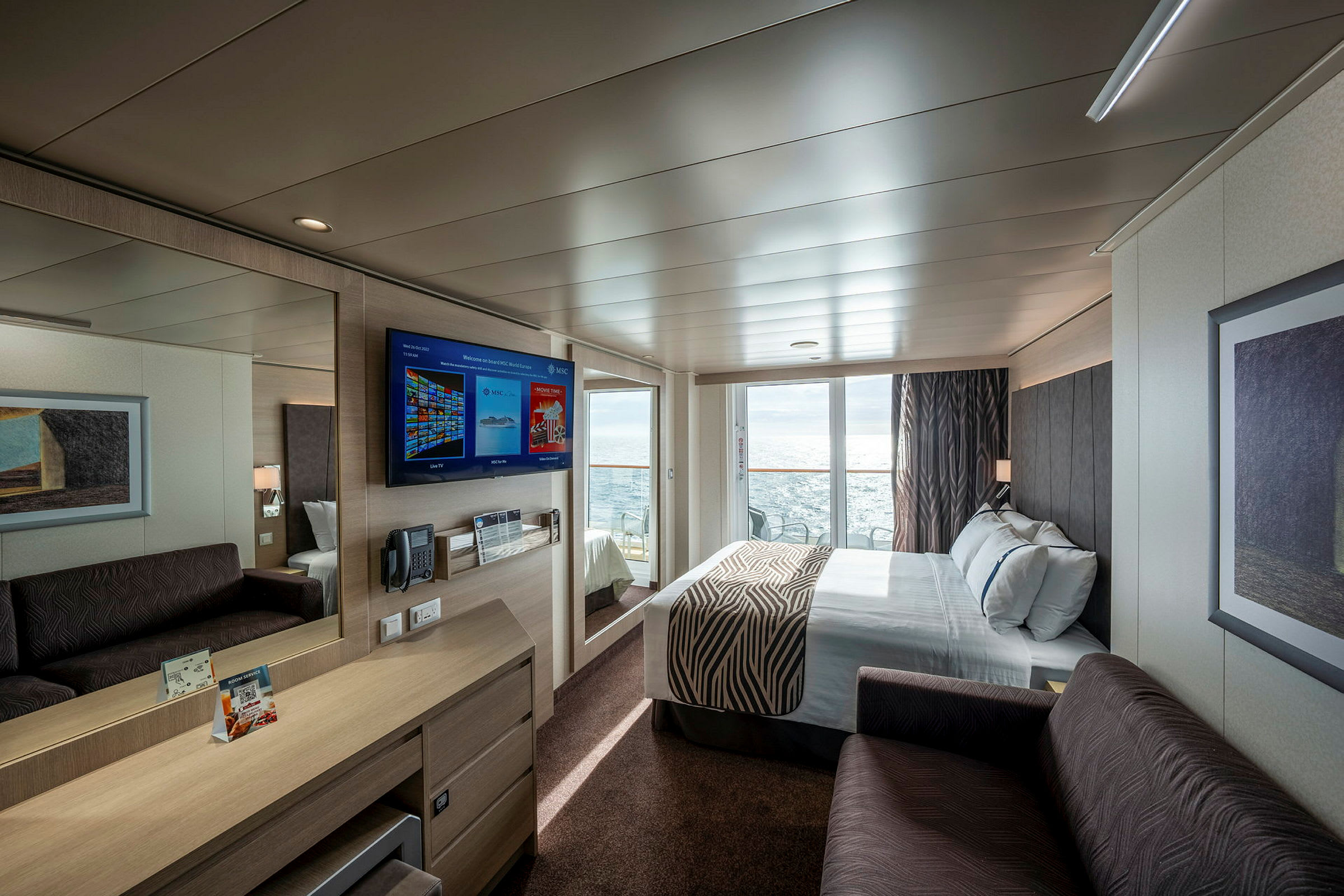 Moderne indrettet hotelværelse med dobbeltseng, sofa, skrivebord og vægmonteret tv. Stor glasdør fører til balkon med havudsigt. (AI-genereret tekst)