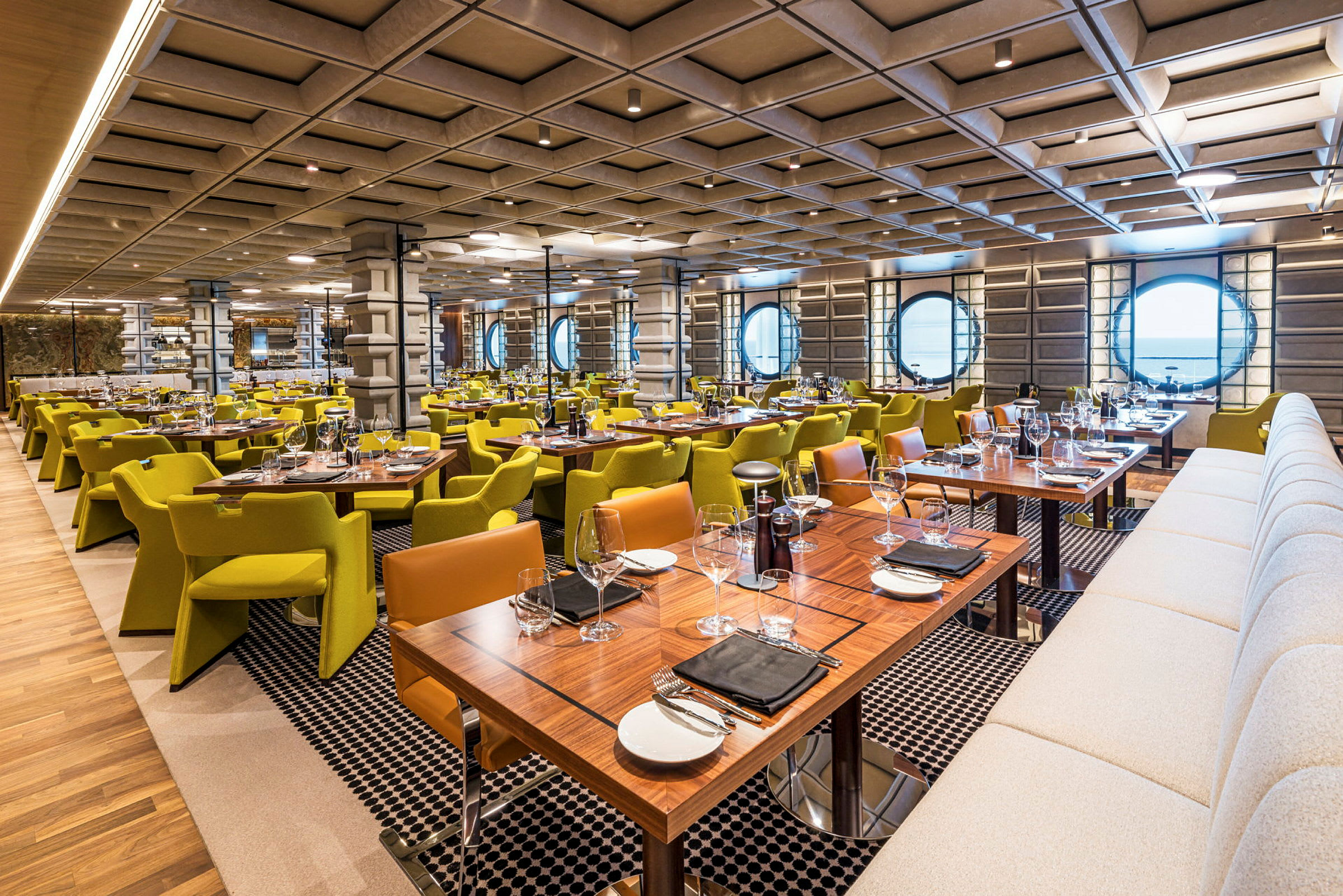 Interiøret i en moderne restaurant med mange dækkede borde, grønne stole og en buet væg med runde vinduer. (AI-genereret tekst)