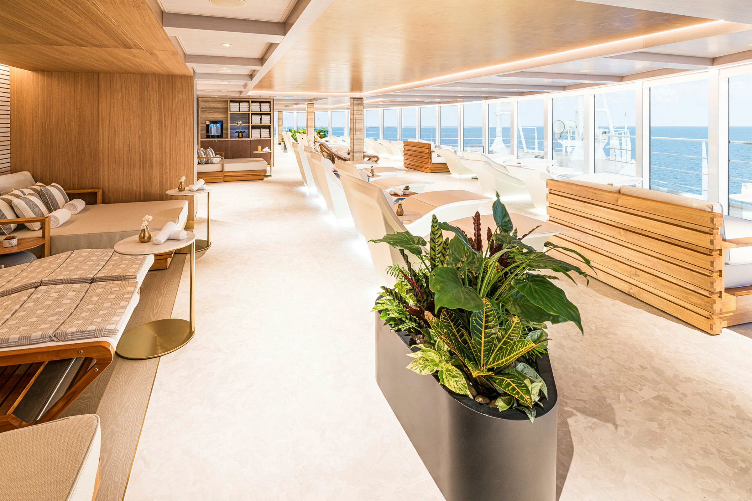 Lys og moderne lounge med liggestole, sofaer og store vinduer med udsigt over havet. Planter i forgrunden. (AI-genereret tekst)
