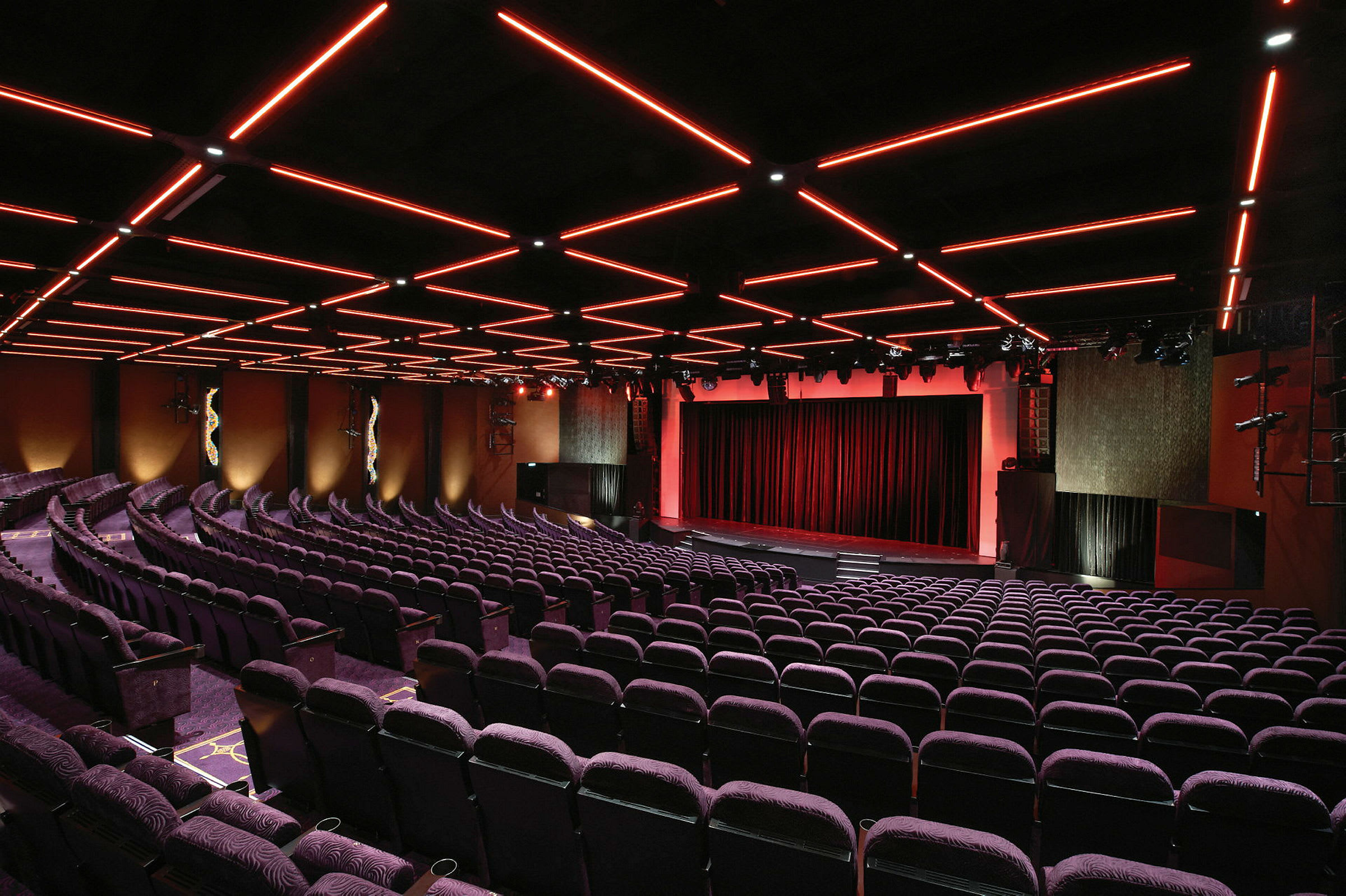 En teatersal med rækker af lilla stole og et oplyst loft med røde linjer. Scenen er beklædt med røde gardiner. (AI-genereret tekst)
