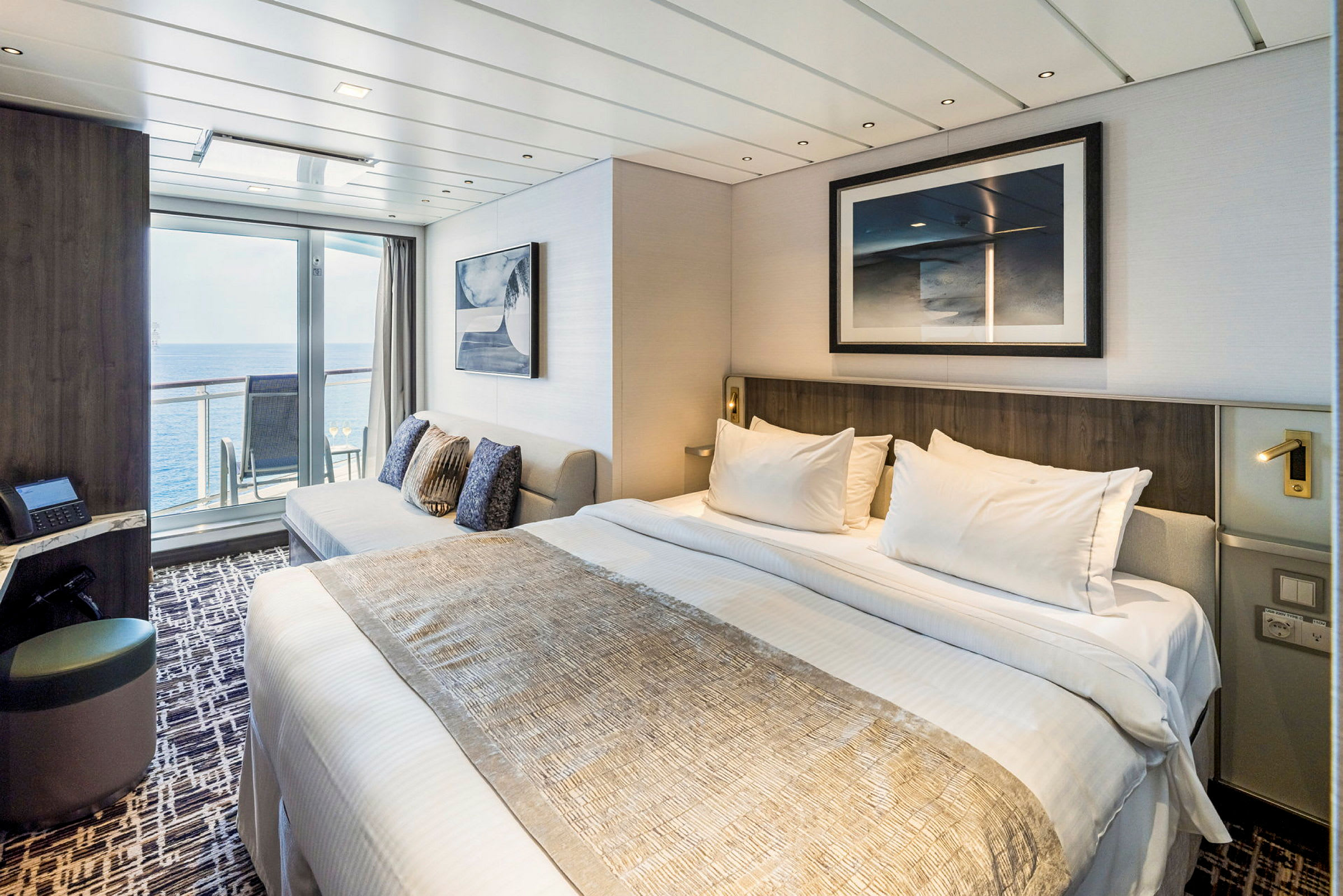Et moderne hotelværelse med en opredt seng, en sofa og glasdøre, der fører ud til en altan med havudsigt. (AI-genereret tekst)