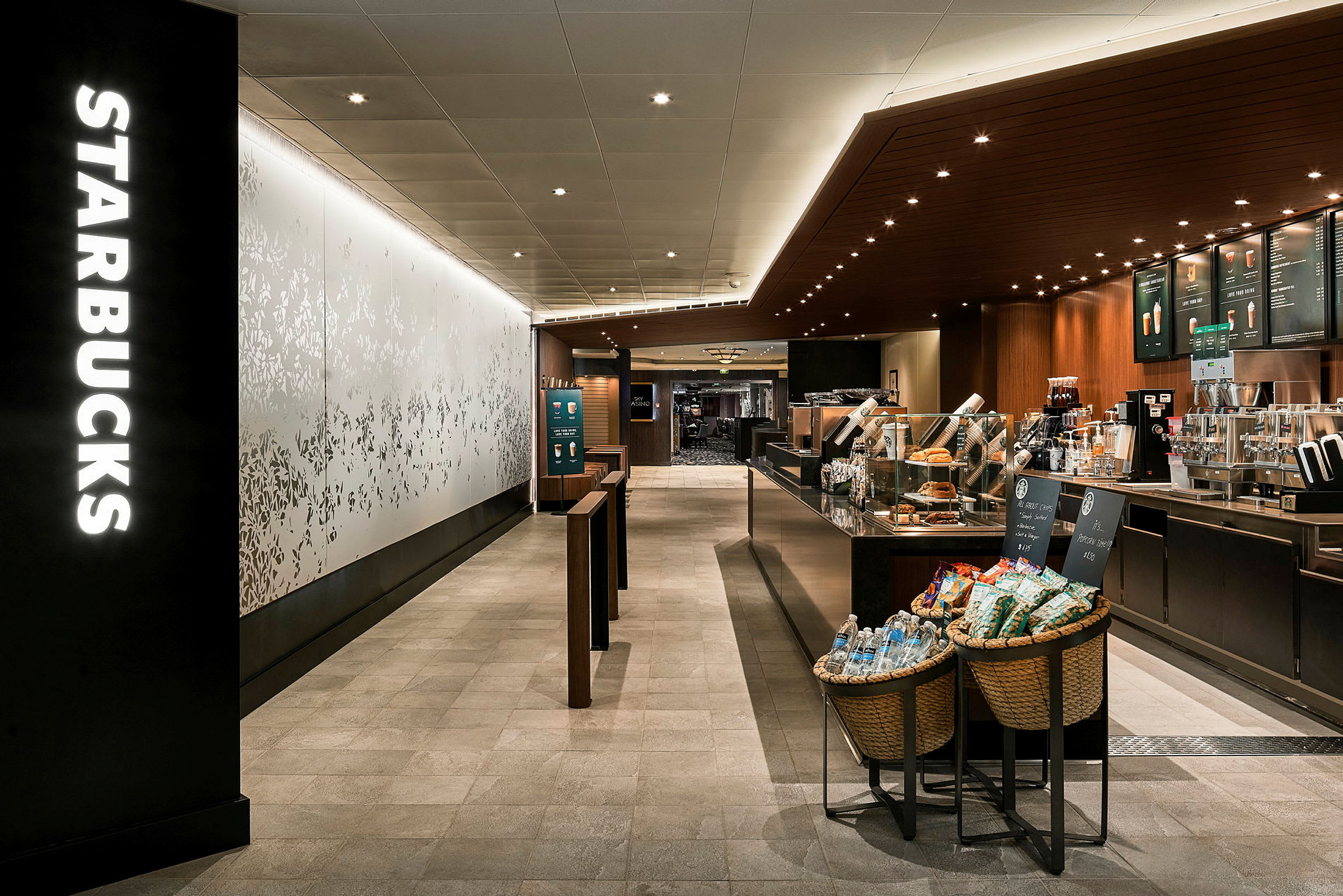 Et interiørbillede af en Starbucks-café med kaffemaskiner, menutavler og varer ved indgangen. (AI-genereret tekst)