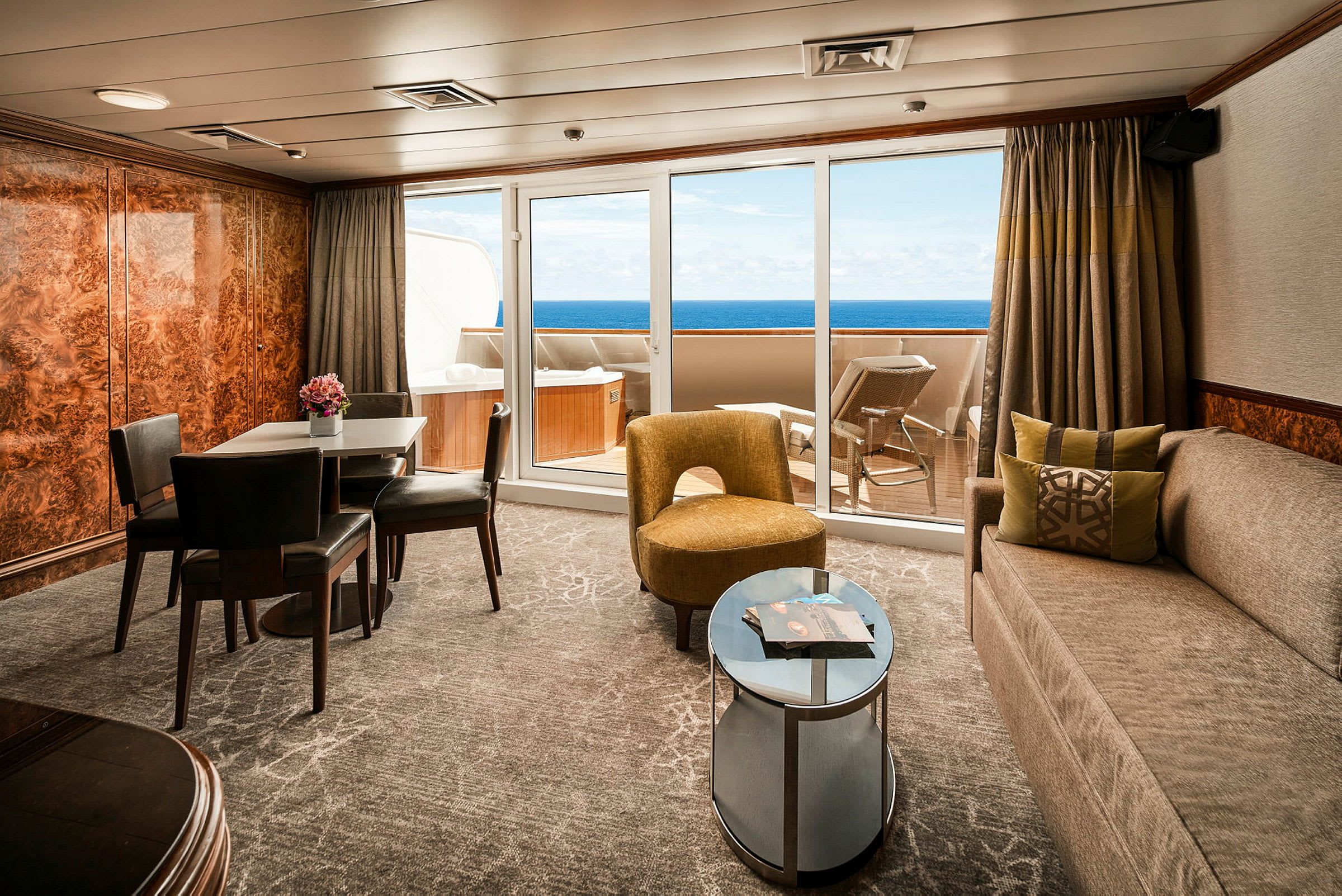 En møbleret stue med spisebord og stole, sofa, lænestol samt udsigt til en balkon med liggestole og havet i baggrunden. (AI-genereret tekst)