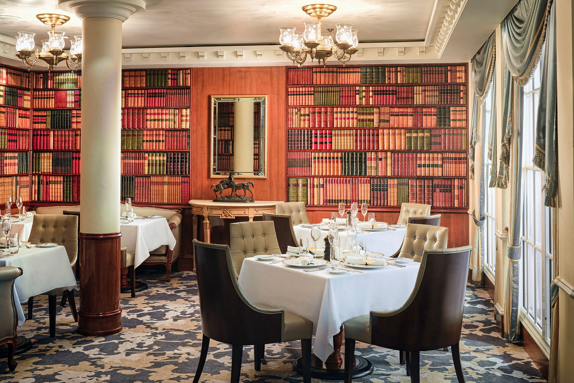En elegant restaurant med borde dækket med hvide duge, omgivet af bogreoler samt stort vindue og rige gardiner. (AI-genereret tekst)