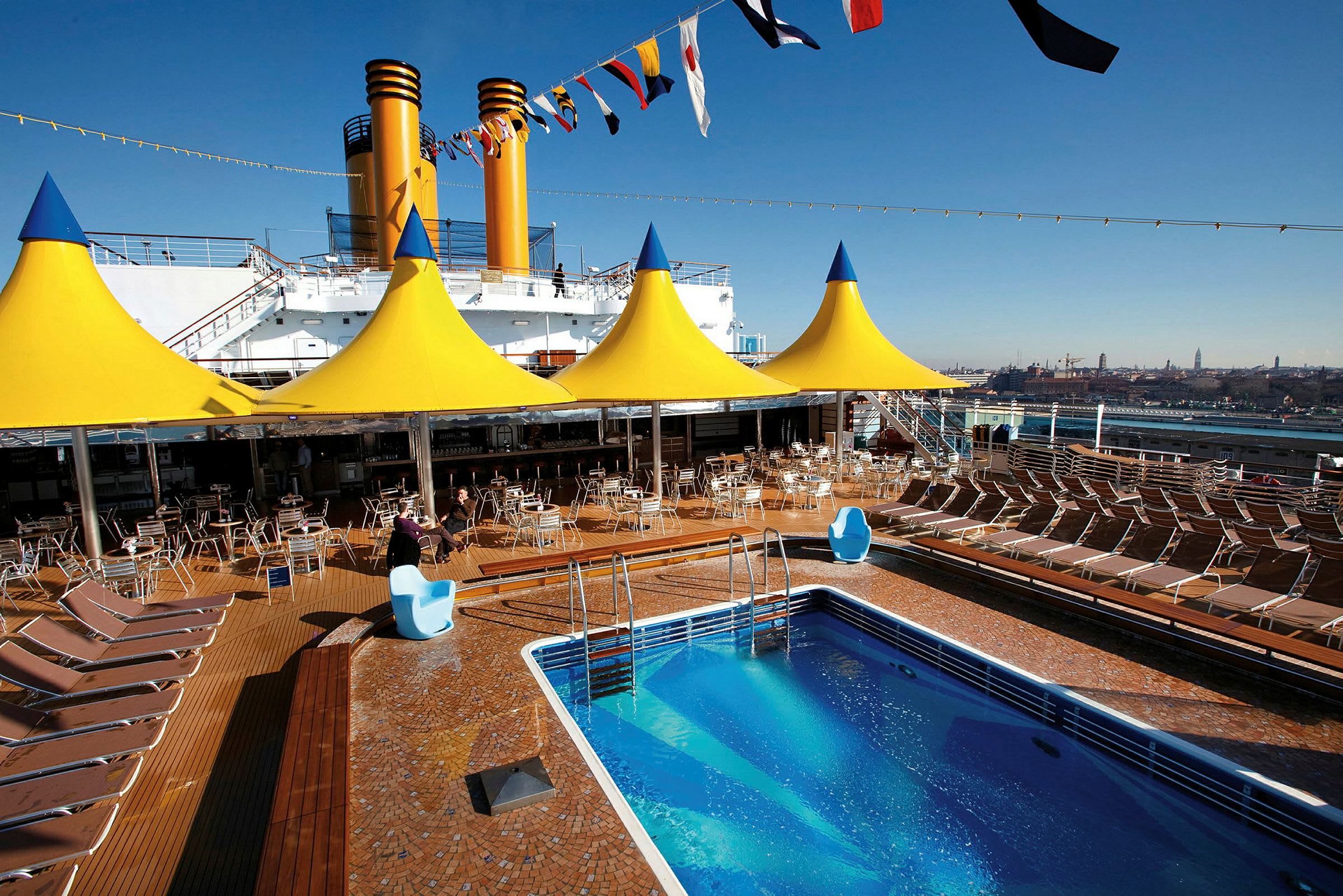 Liggestole ved en swimmingpool på dækket af et krydstogtskib, med gule teltlignende konstruktioner ovenover. (AI-genereret tekst)