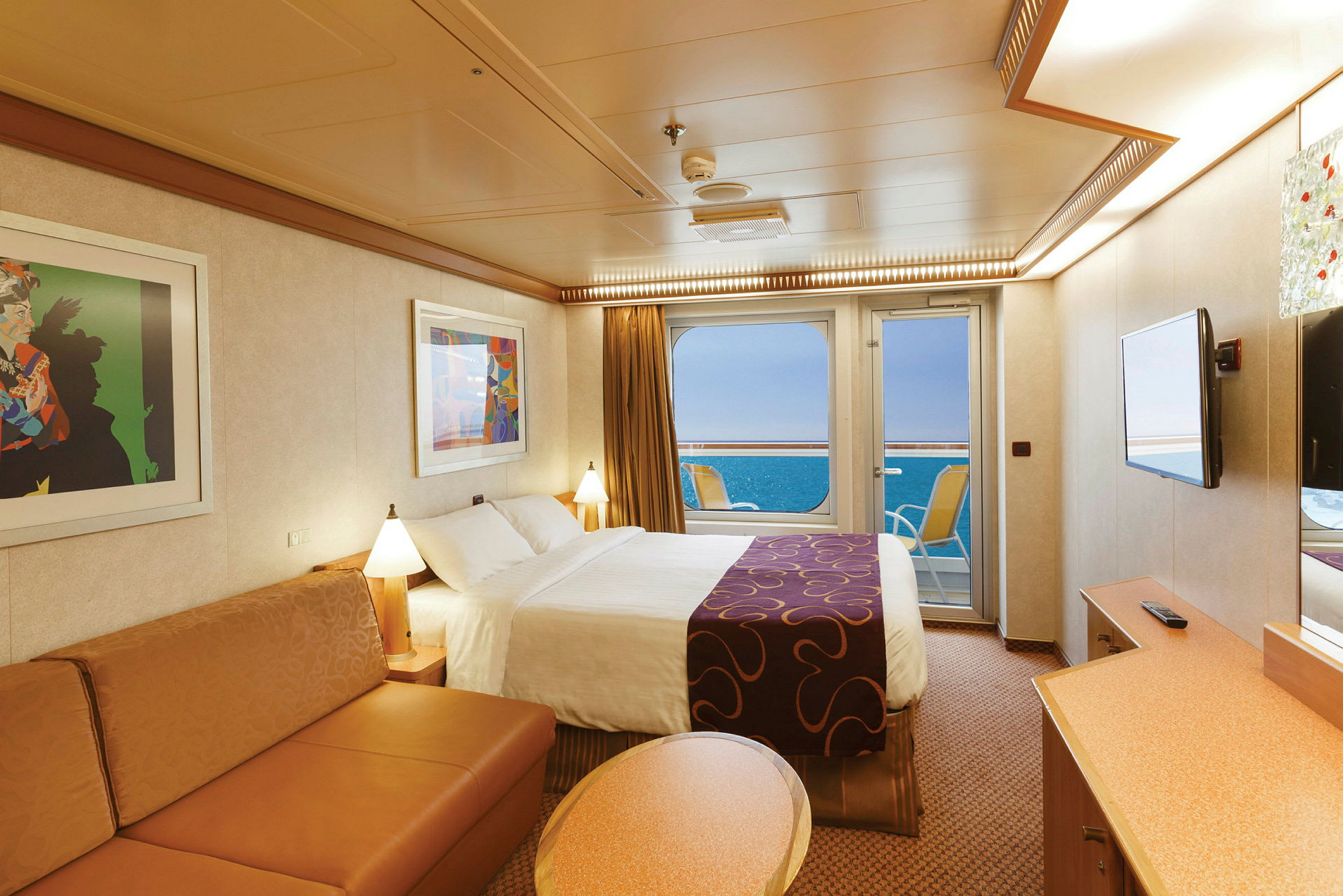Et hotelværelse med seng, sofa, to malerier og en glasdør ud mod havet. (AI-genereret tekst)