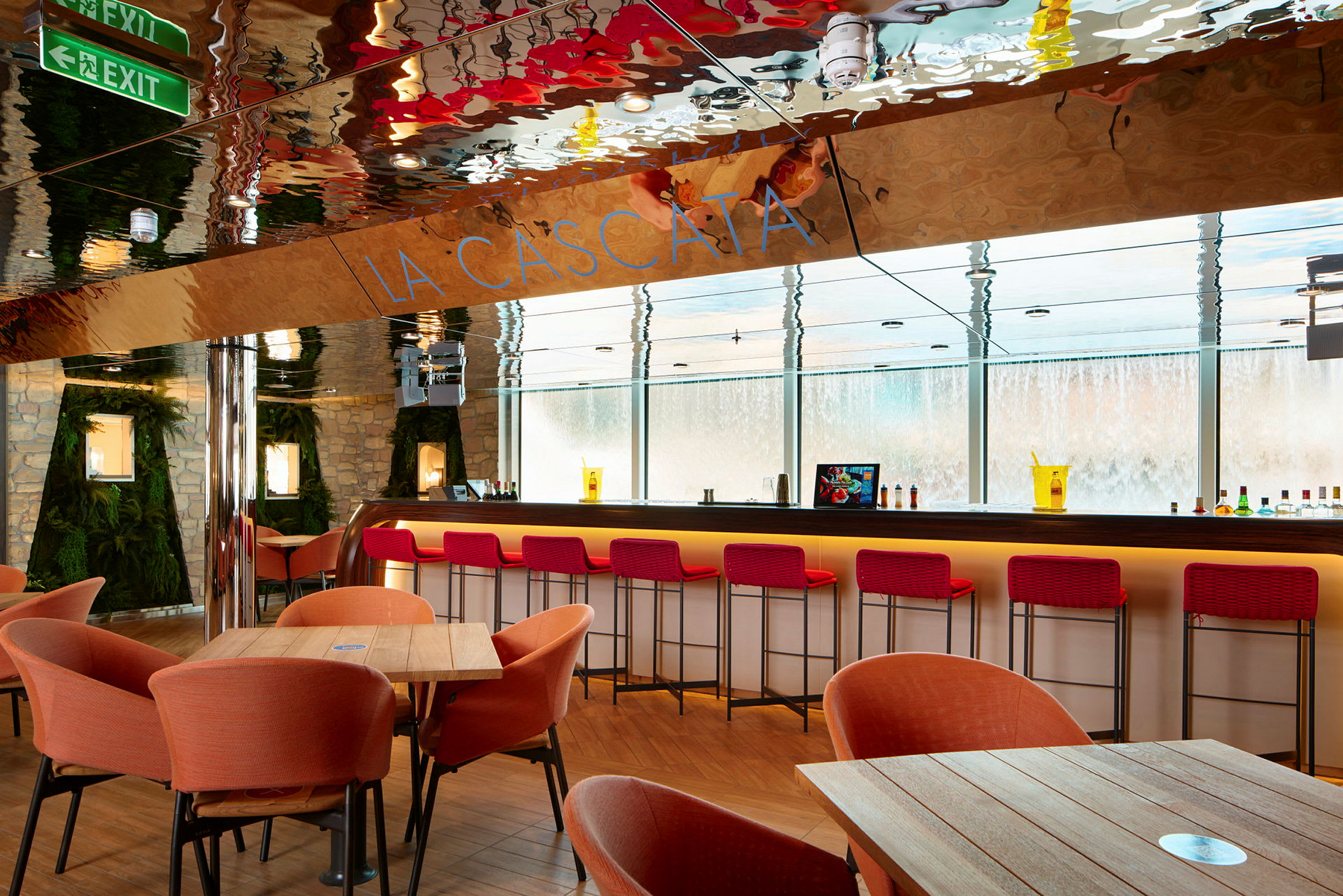 En moderne bar med røde barstole, orange lænestole og træborde. Væggene er dekoreret med sten og grønt. Glasvinduer viser en vandinstallation udenfor. (AI-genereret tekst)