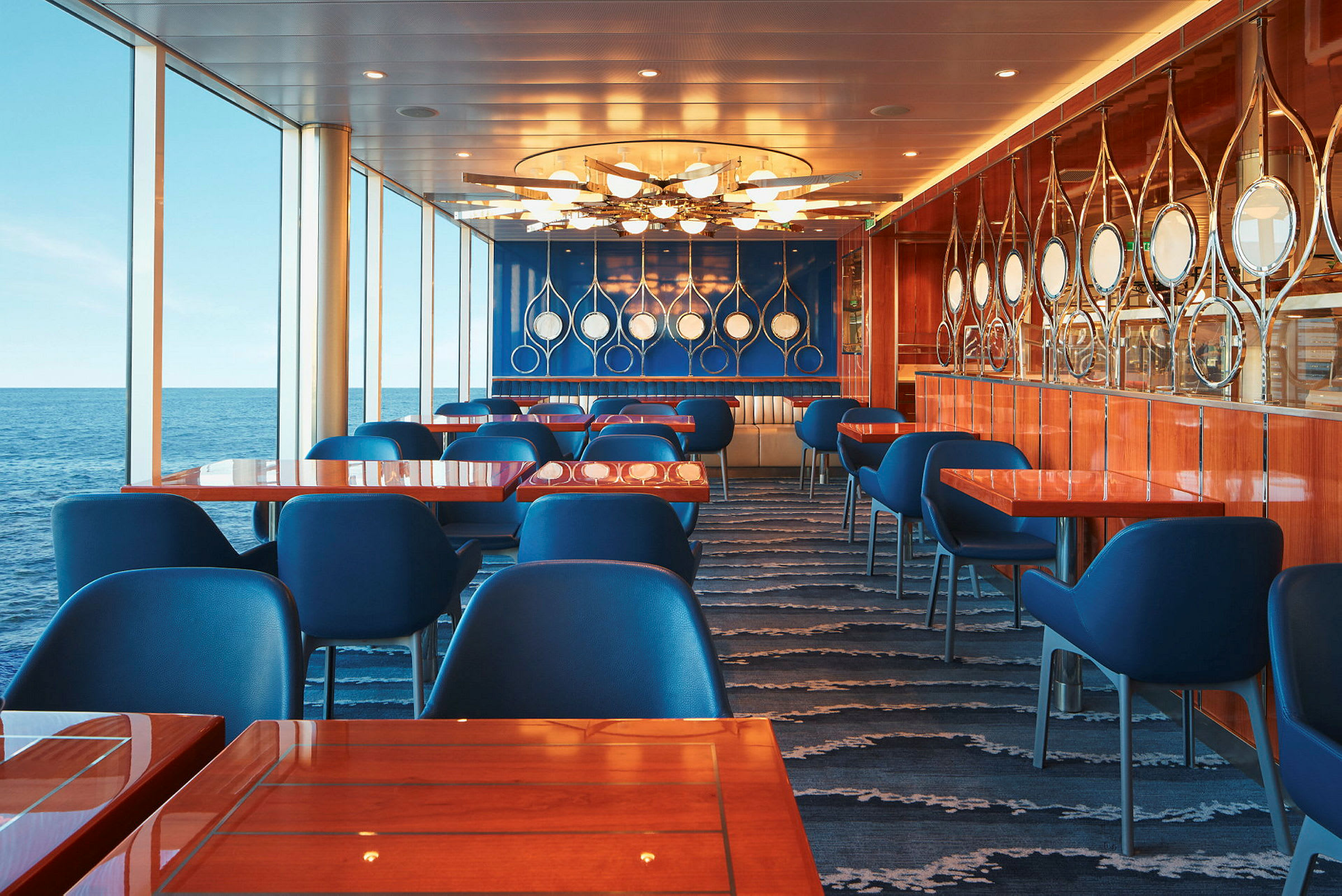 Restaurant med blå stole, borde med træoverflade og panoramavinduer mod havet. (AI-genereret tekst)