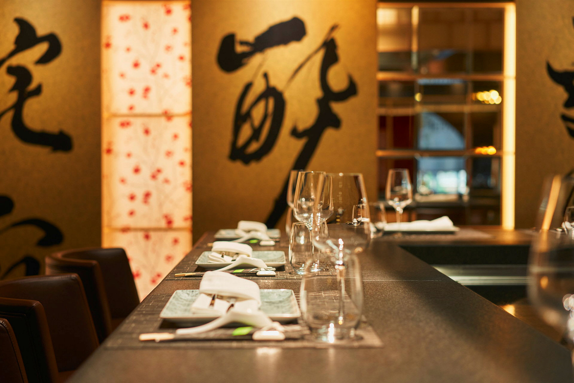 Et dækket bord på en restaurant, dekoreret med glas, porcelæn og bestik. I baggrunden ses dekorationer med asiatiske motiver. (AI-genereret tekst)
