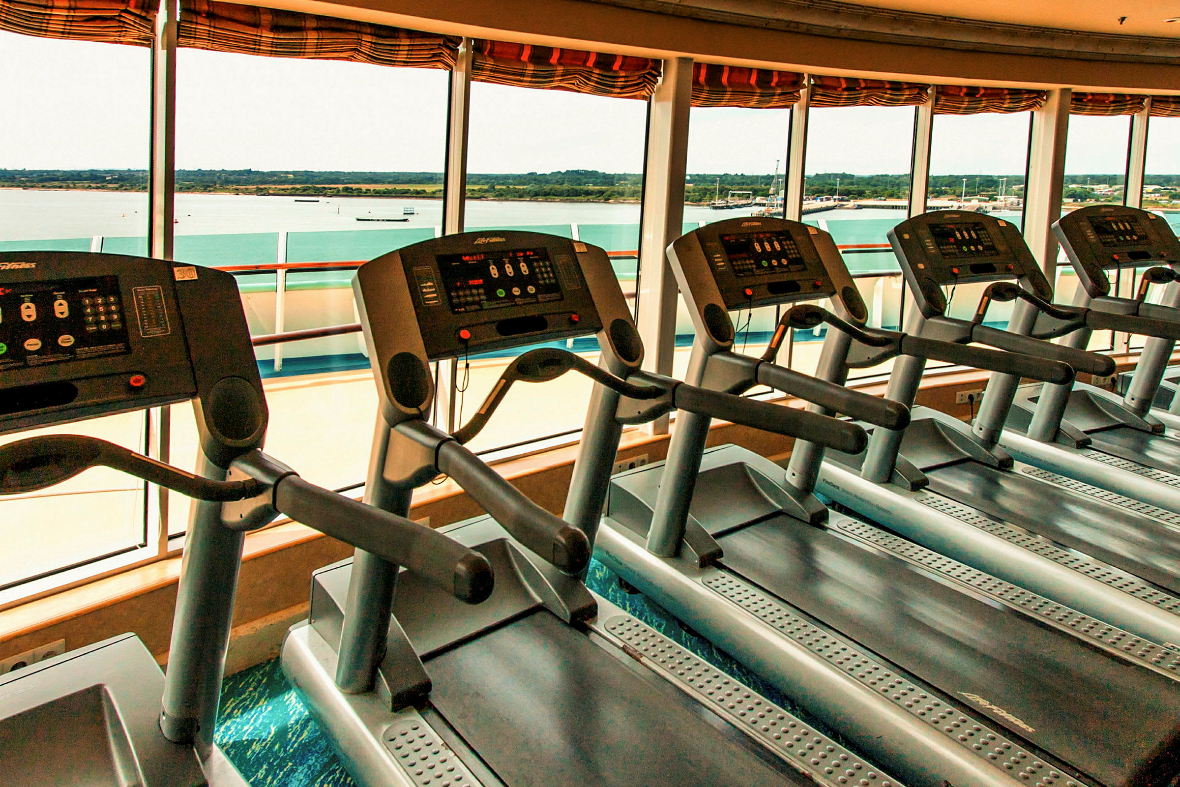 Løbebånd i et fitnesscenter med udsigt over vandet gennem store vinduer. (AI-genereret tekst)