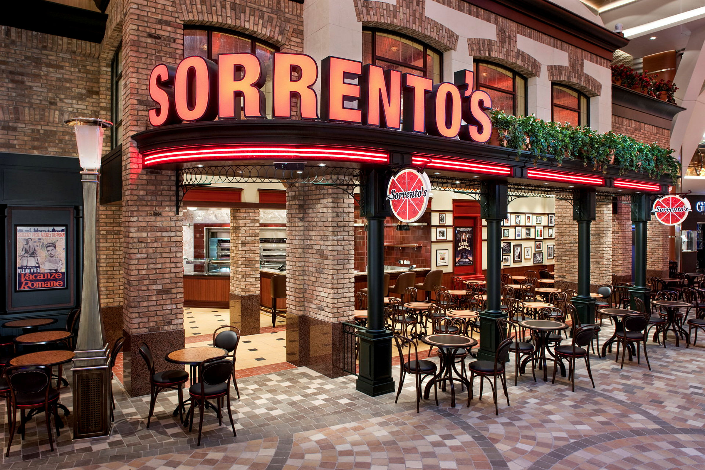 Eksteriør af en restaurant ved navn "Sorrento's". Bygningen har murstensvægge og røde neonskilte. Foran restauranten findes små borde og stole. (AI-genereret tekst)
