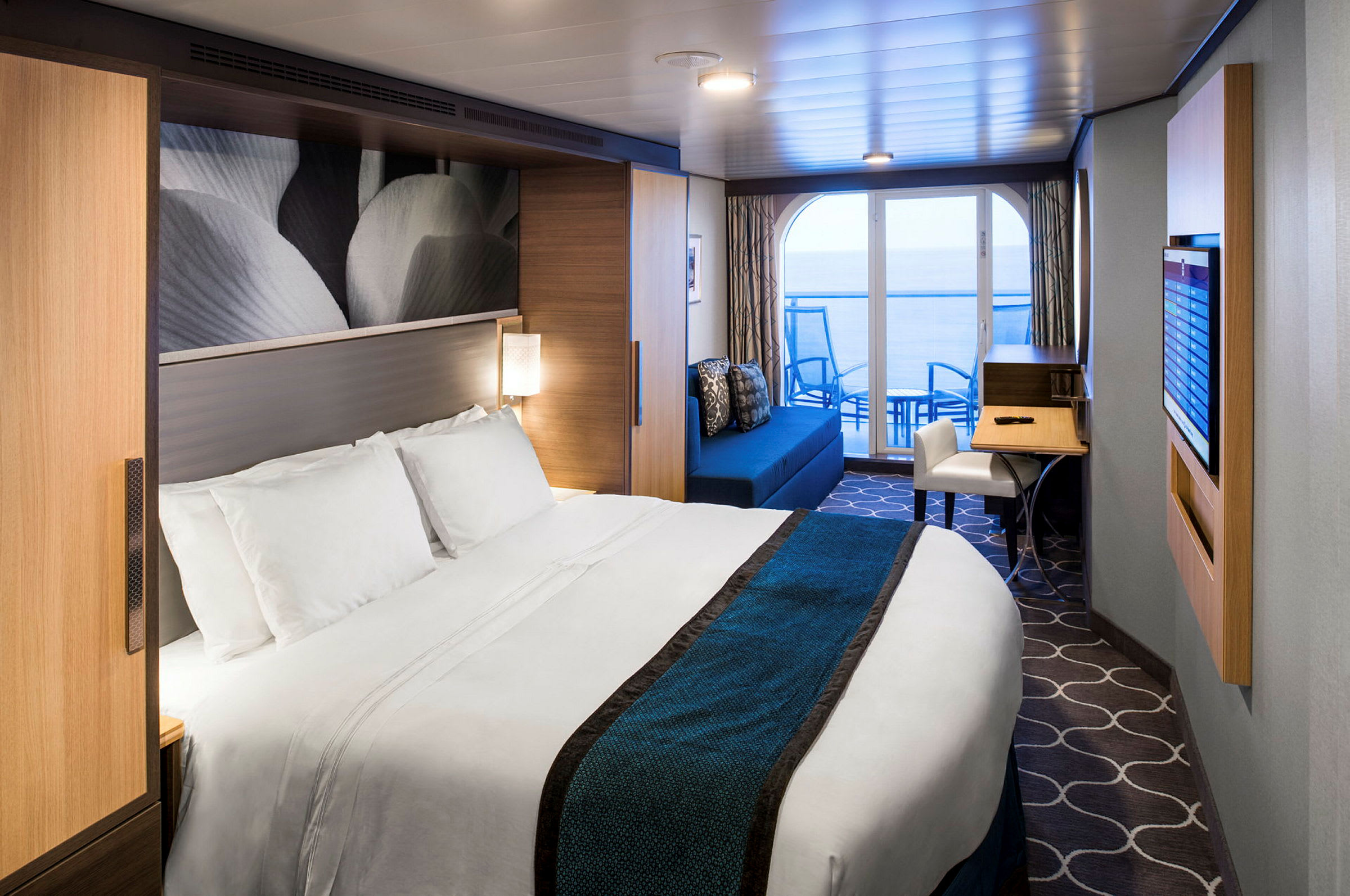 Et moderne hotelværelse med en stor seng, dekoreret med hvide og blå sengetøj. Værelset har en siddegruppe og skrivebord samt en altan med udsigt til havet. (AI-genereret tekst)