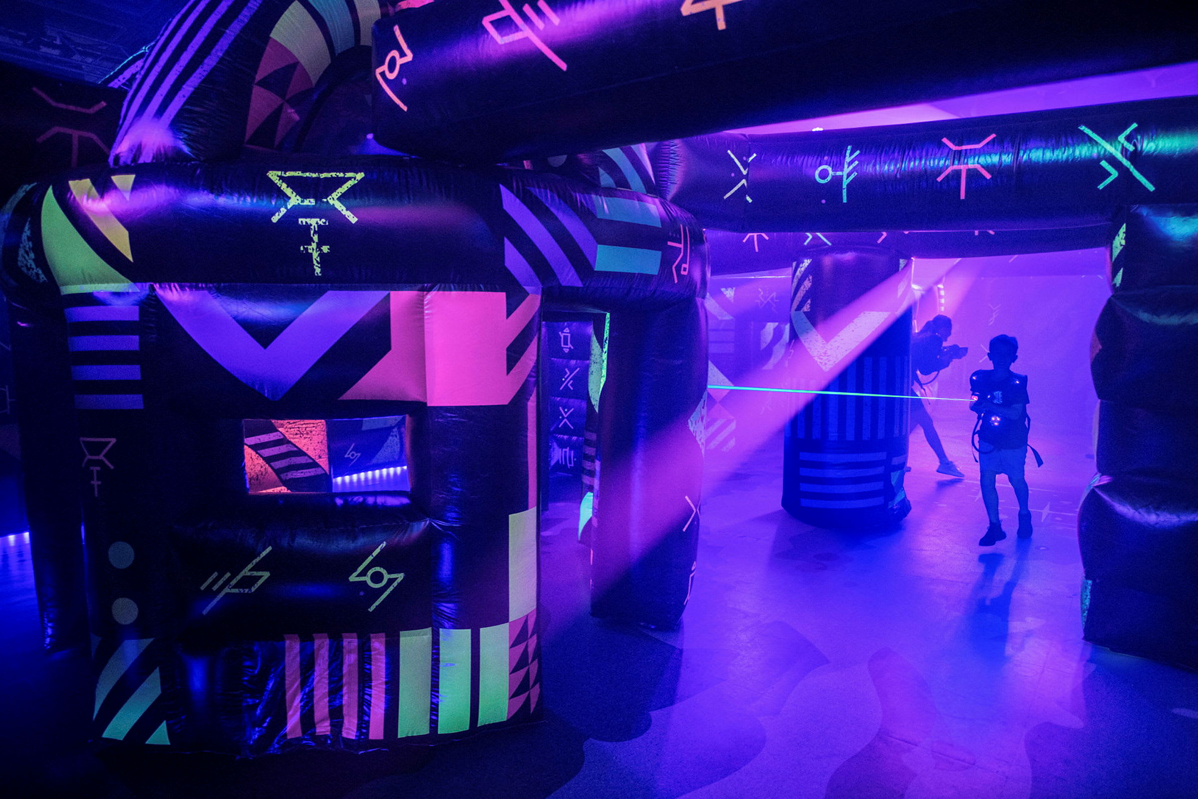 Et neonbelyst indendørsmiljø med oppustelige konstruktioner og personer, der spiller laser tag. (AI-genereret tekst)