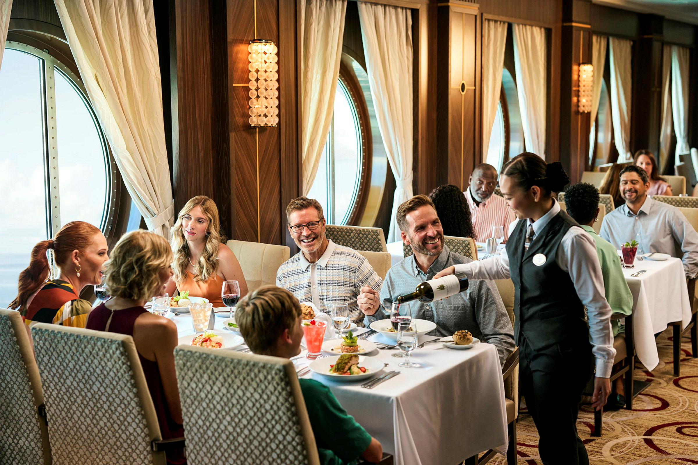 En elegant restaurant med et vindue med udsigt over havet, hvor mennesker nyder et måltid, og en tjener serverer vin. (AI-genereret tekst)